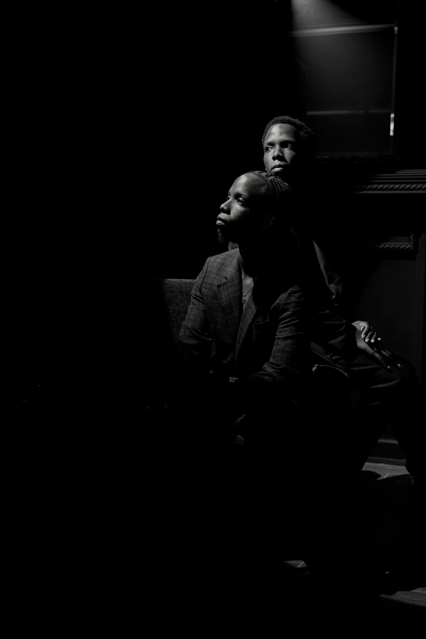 Two people sitting in a dark room with minimal lighting, one in the foreground and one in the background, both looking to the side.
