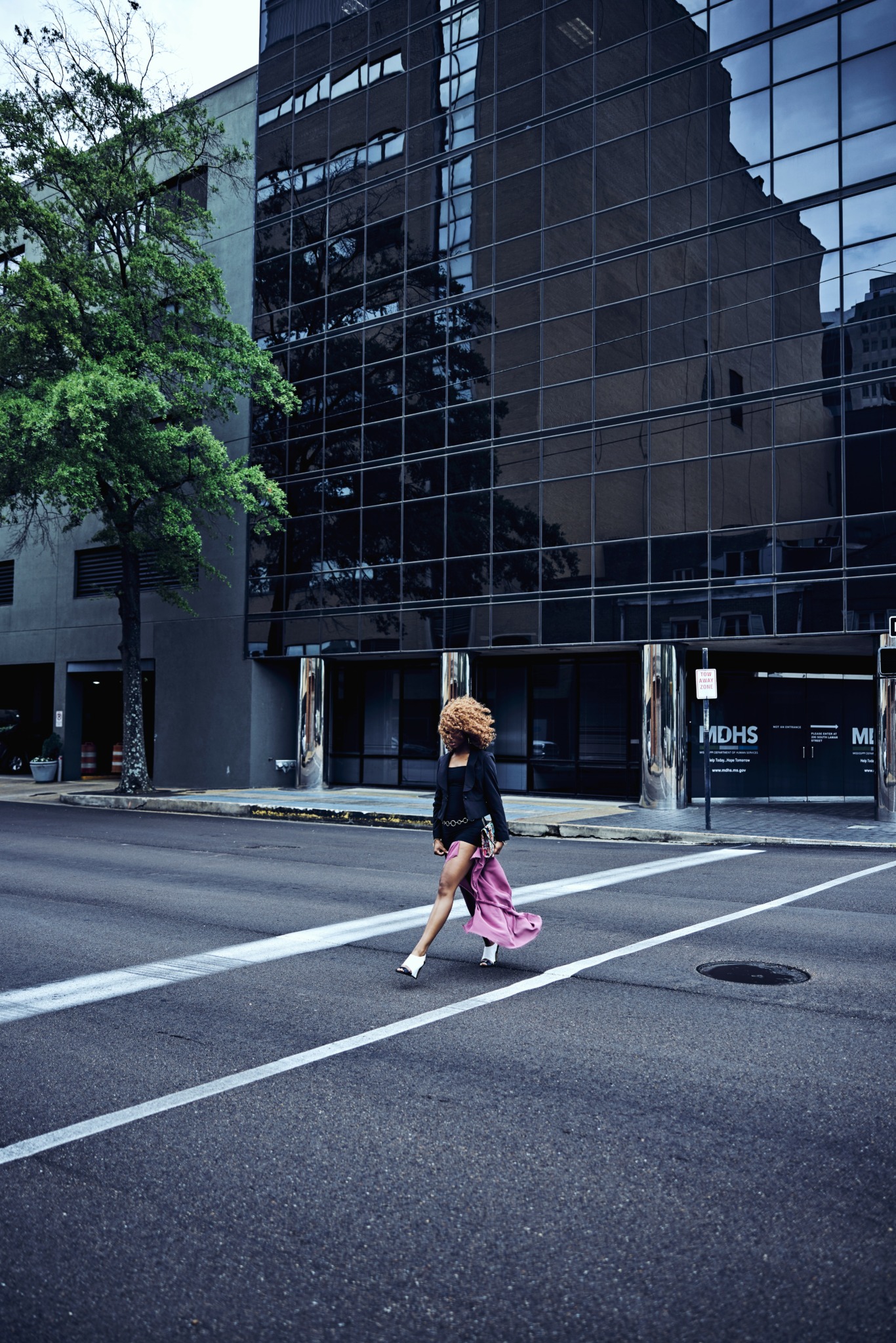 Person crossing street with pink bag in front of a reflective glass building, trees on the sidewalk.