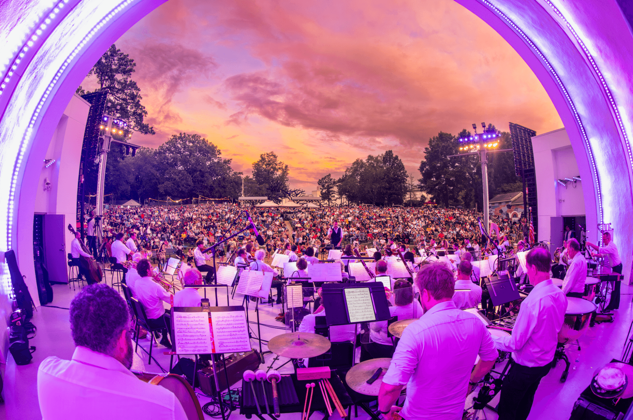 Orchestra performing outdoors at sunset with a large audience and trees in the background.