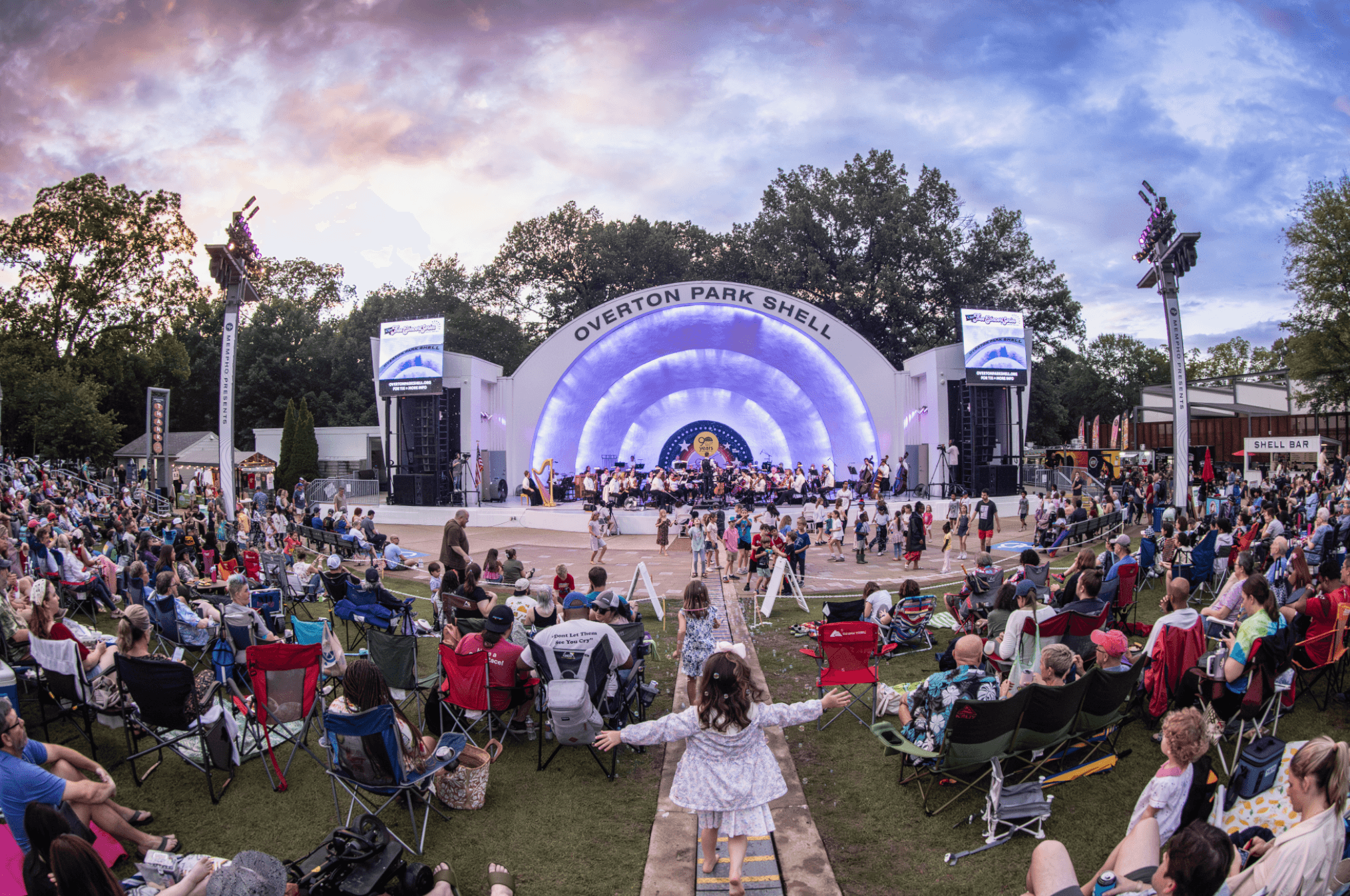 Large outdoor concert with a stage, audience seated on grass, and a girl in a white dress watching.