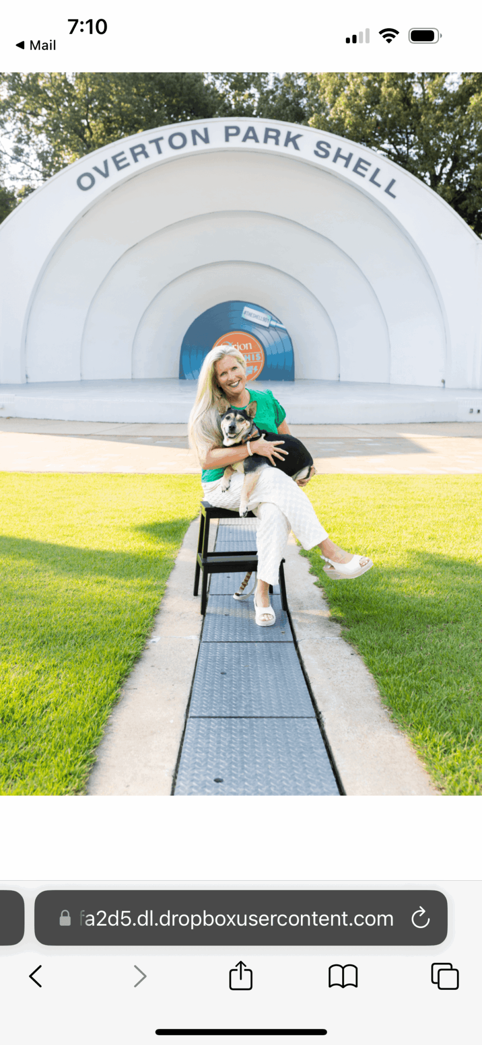 Person sitting on a bench in front of a large white shell structure with 'Overton Park Shell' written on it, holding a green bag.