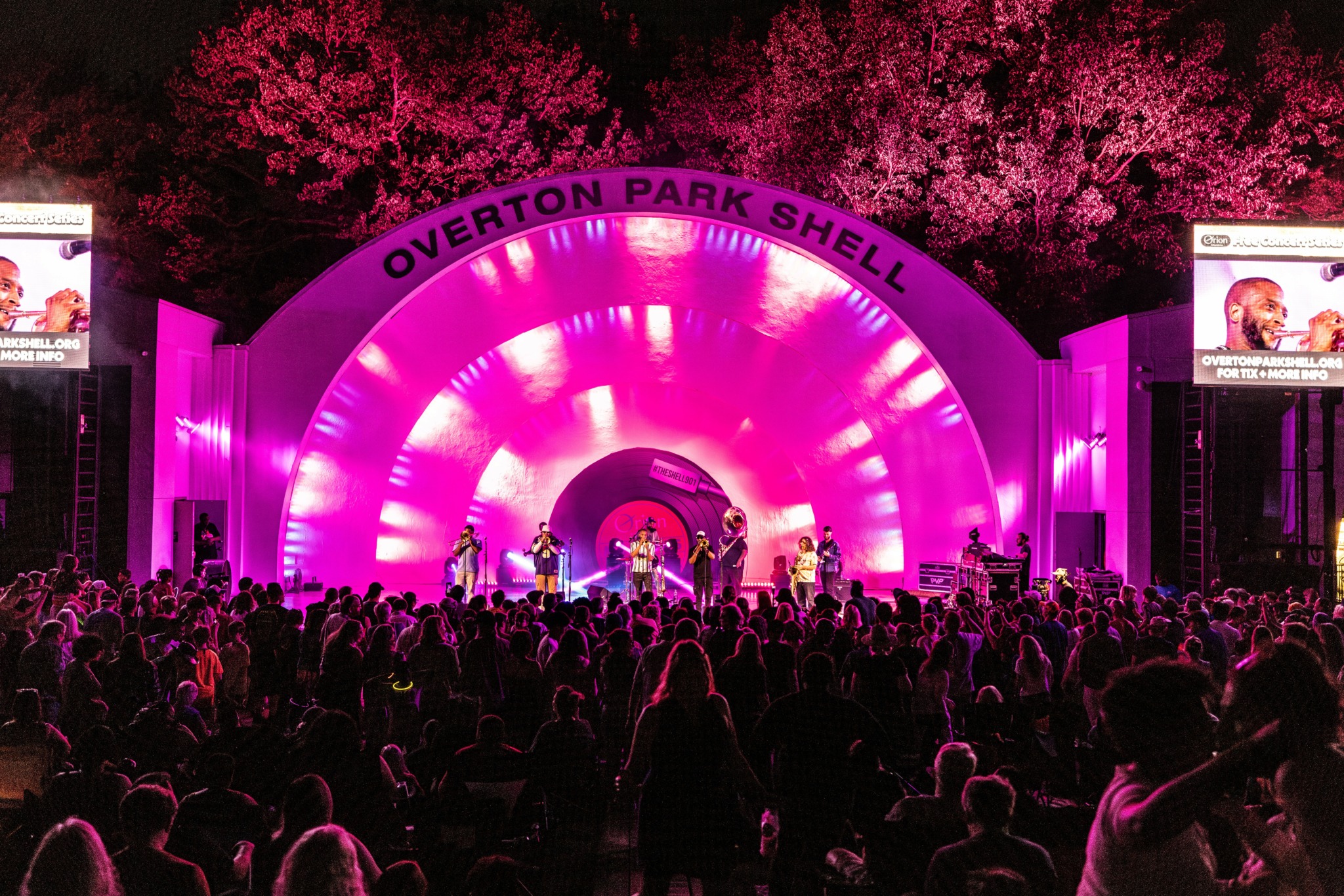 Outdoor concert stage illuminated with pink lights, crowd watching, trees in background, screens on sides, at Overton Park Shell.