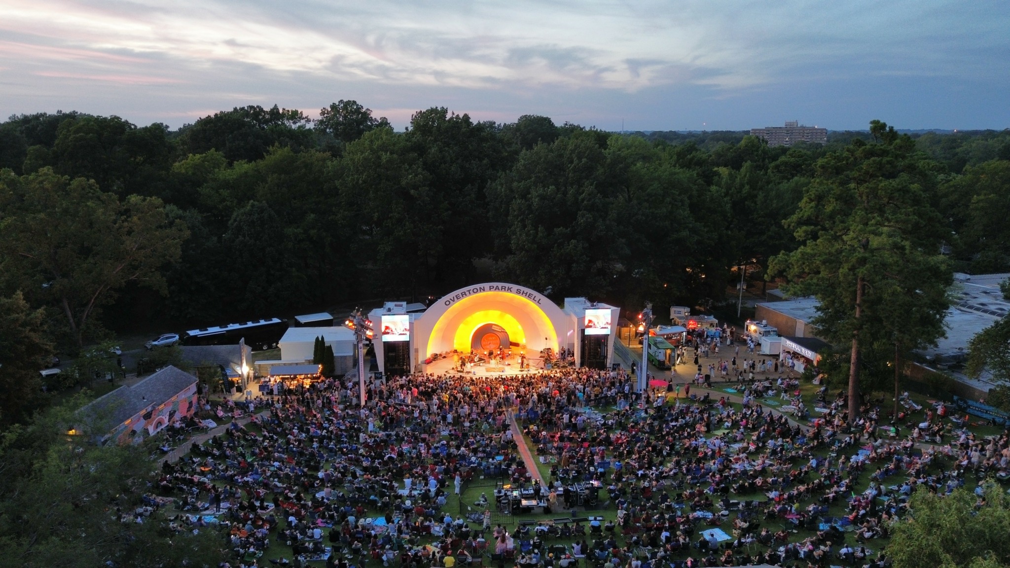 Large outdoor concert with a stage, crowd, and trees in the background during dusk.
