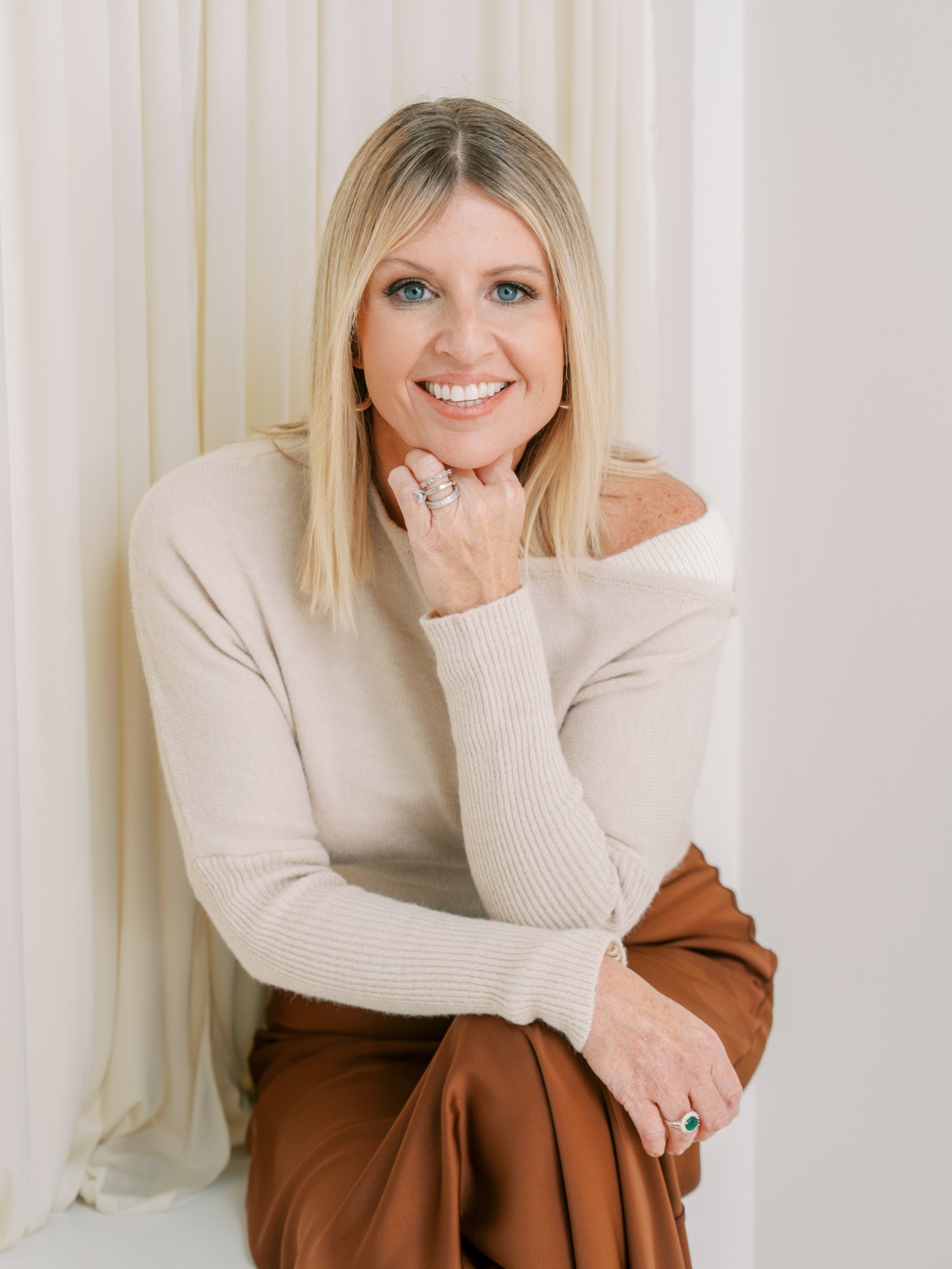 Woman with blonde hair smiling, wearing a cream sweater and brown pants, sitting near a white wall and curtain.