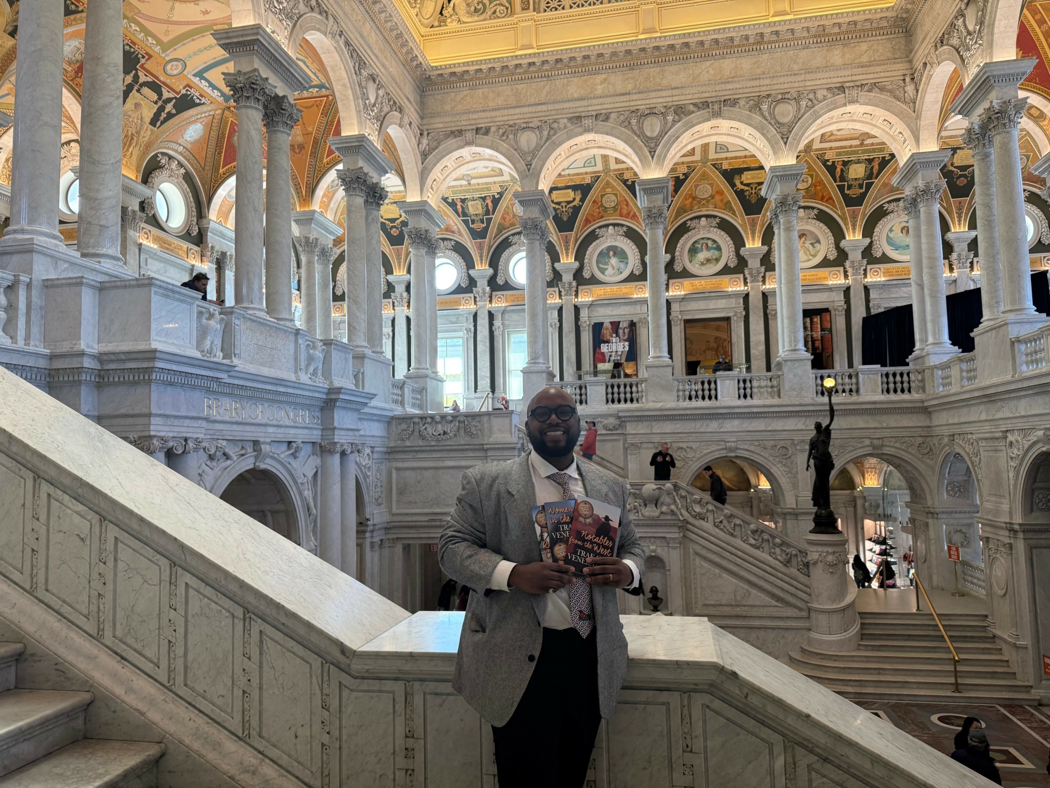 Man in a suit holding a book inside a grand, ornate building with arches and columns.