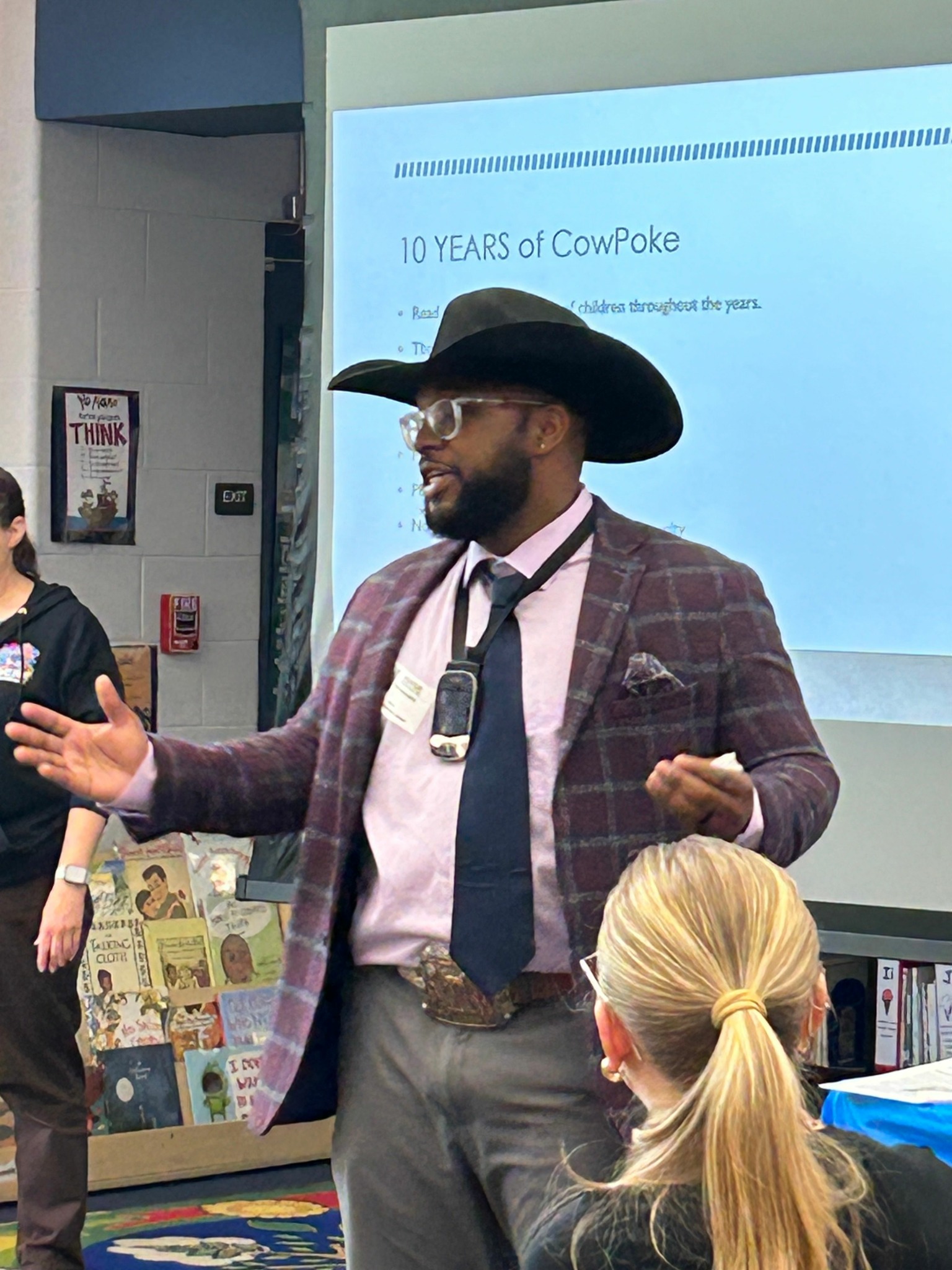 Person in a plaid jacket and hat speaking to a group in a classroom, with a woman listening nearby.
