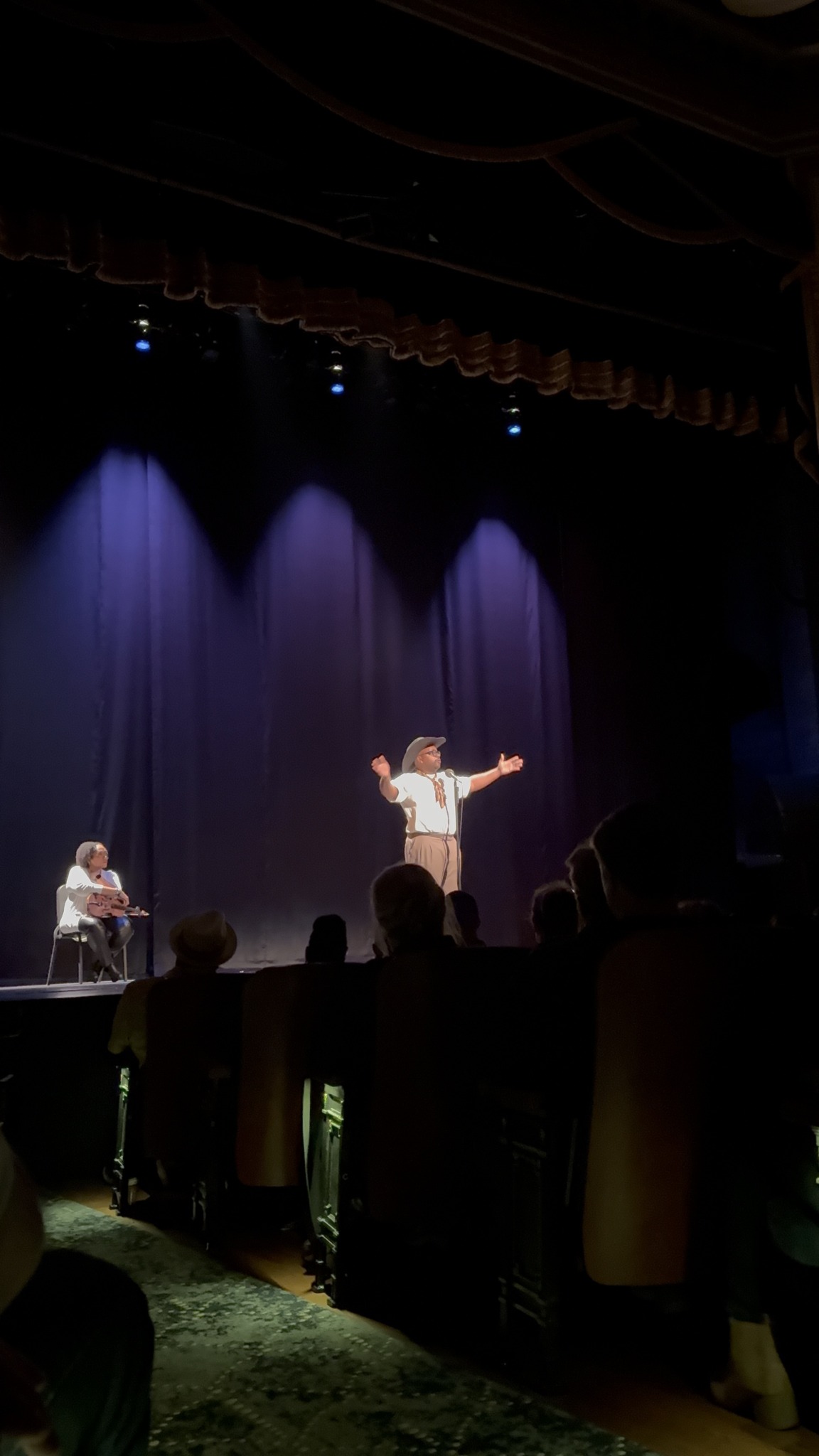 Performer on stage with arms outstretched, seated musician nearby, audience watching in a theater with purple lighting.