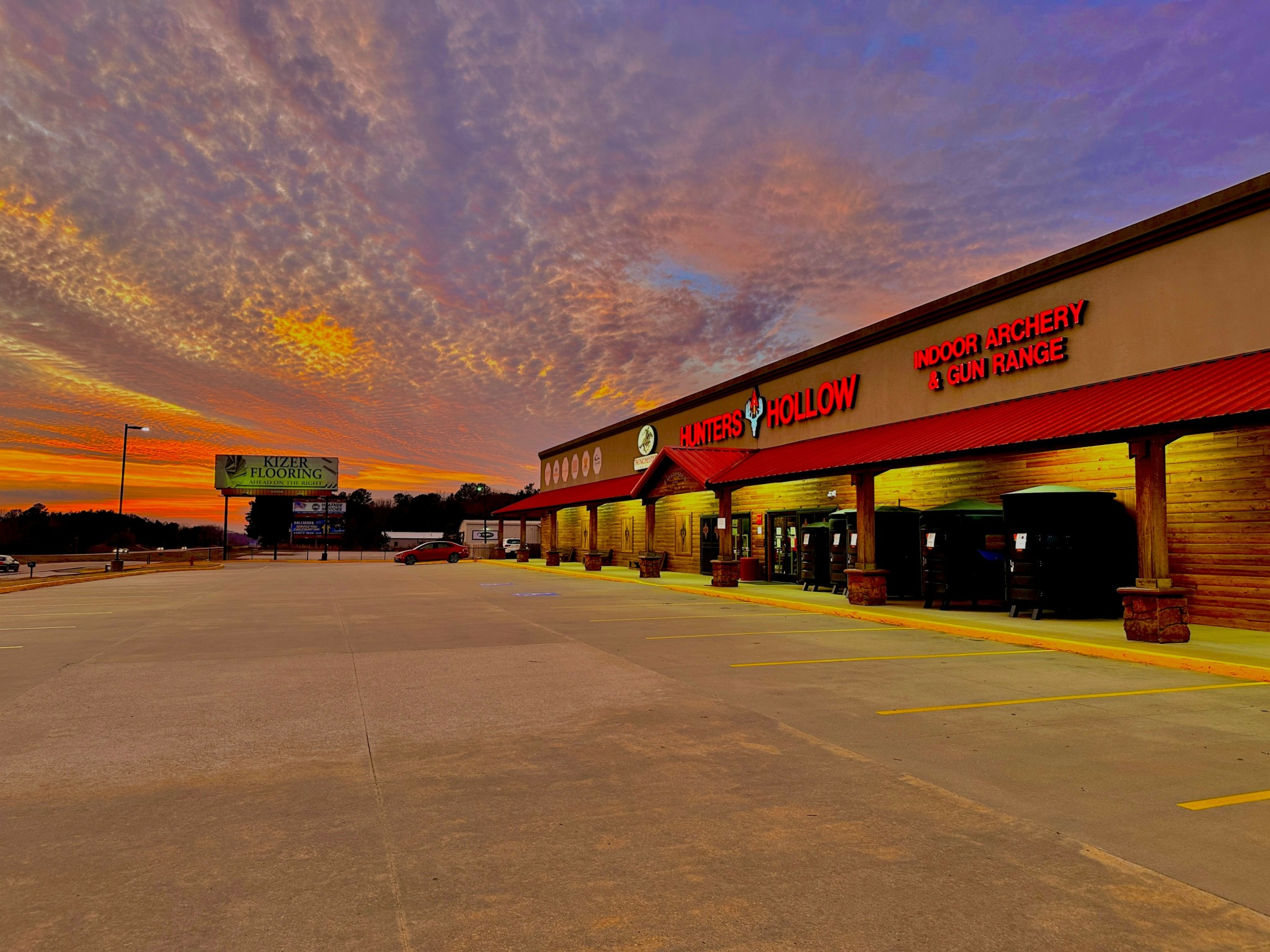 Empty parking lot in front of a shopping center during sunset with colorful sky, storefronts, and parking spaces.