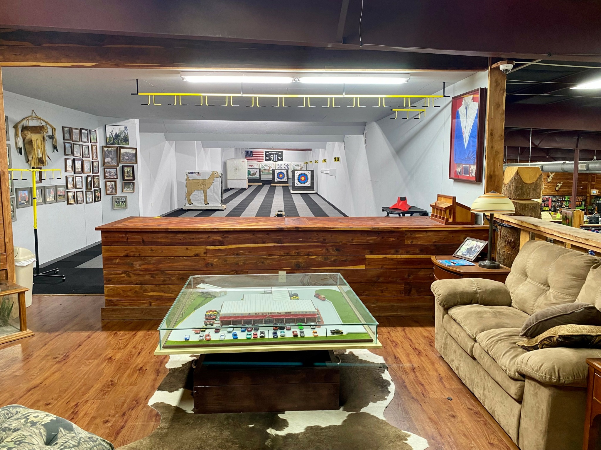 Indoor shooting range with targets, wooden flooring, and seating area, including a glass display table and a beige sofa.