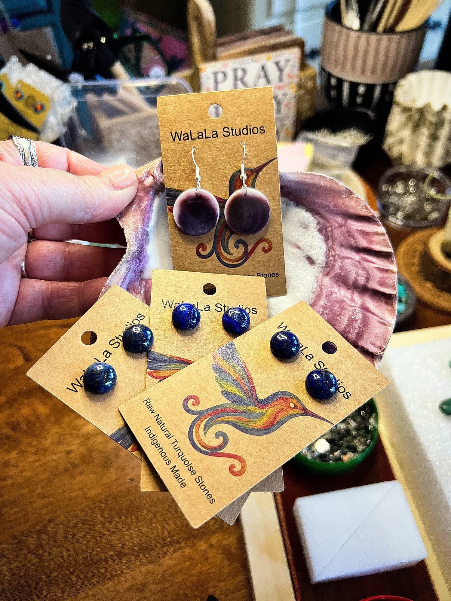 Hand holding earrings and three cards with jewelry, background shows various craft supplies and decorative items.