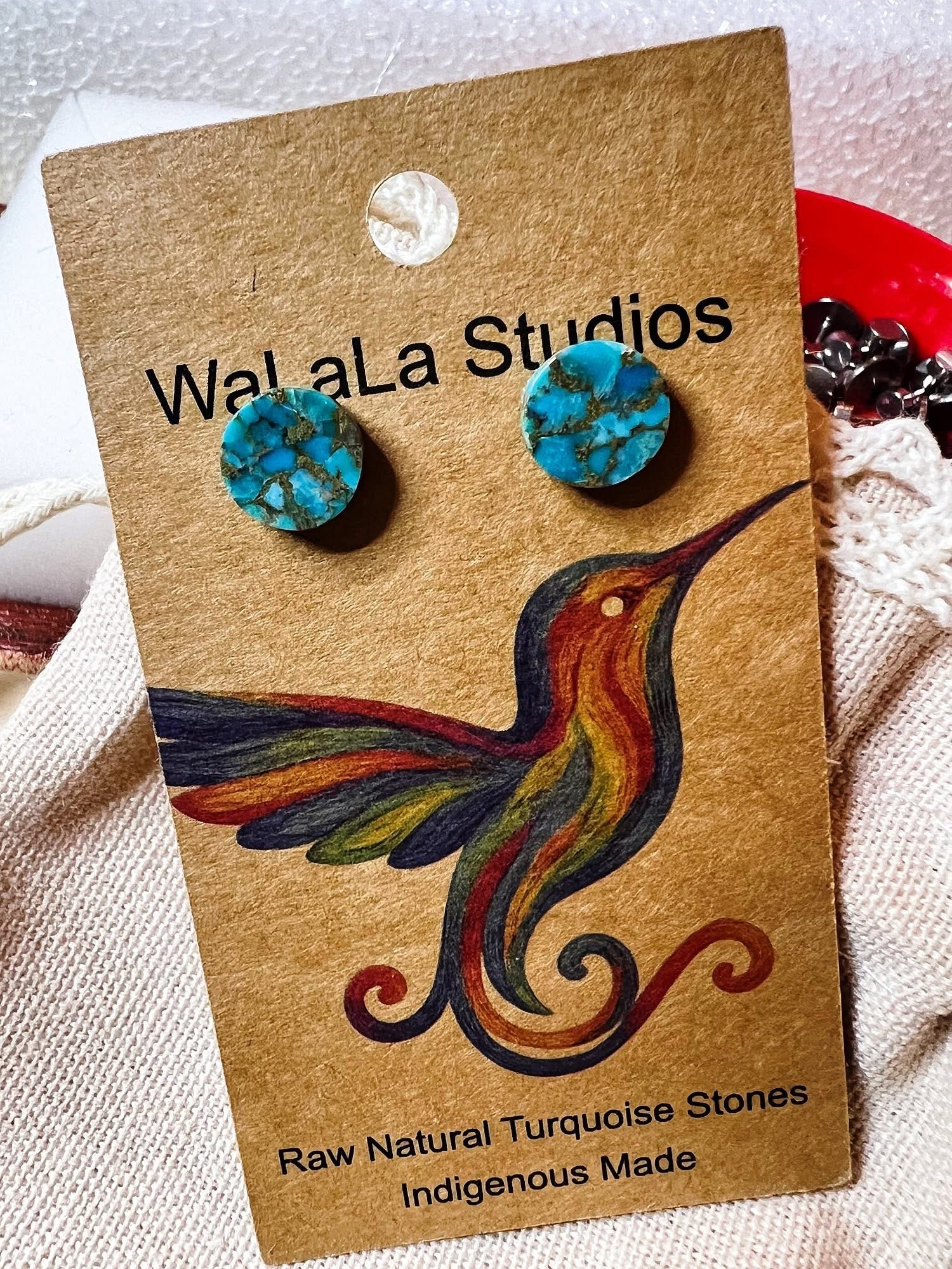 Pair of turquoise and black earrings on a card with a colorful hummingbird illustration, text about natural stones, and a hole at top.
