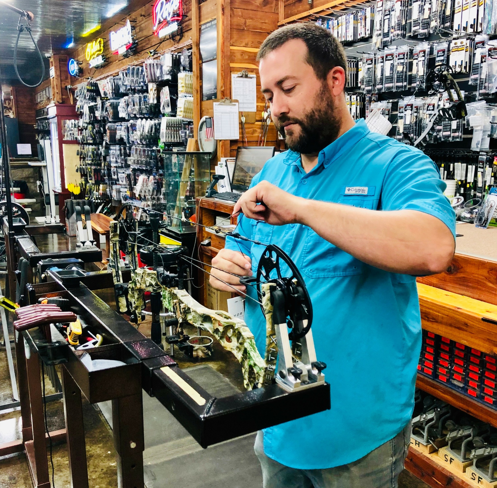Man in blue shirt examining fishing reels in store, shelves with tools and accessories in background.