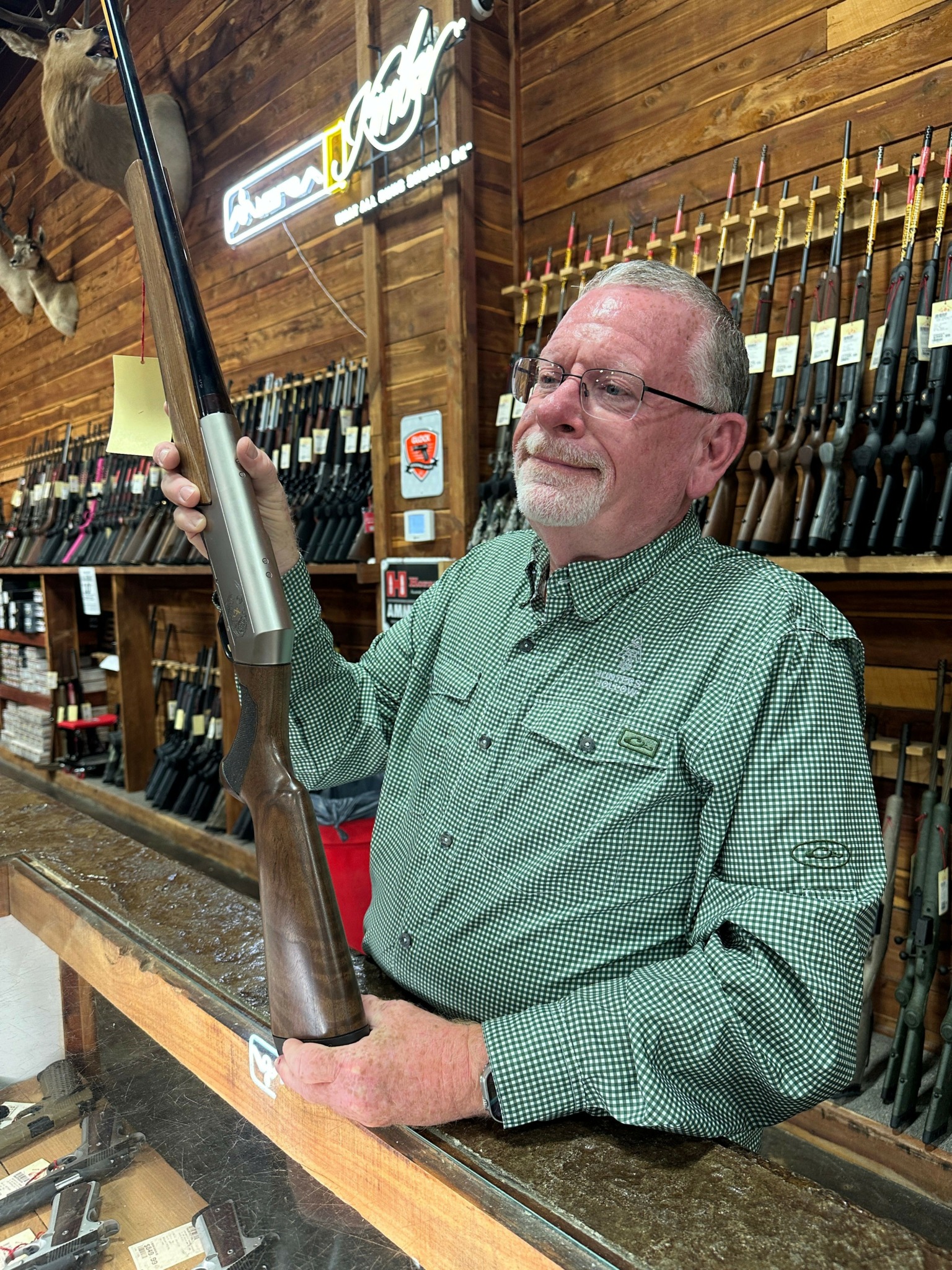 Man with glasses and gray hair in a green shirt at a wooden counter in a wine shop, holding a rifle.
