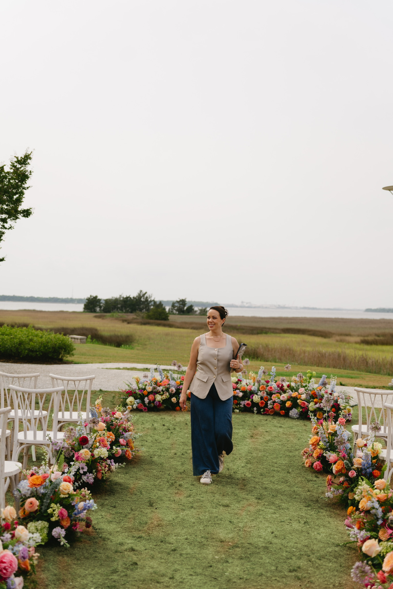 Woman walking down a grassy aisle decorated with colorful flowers, outdoors with a cloudy sky and open landscape in background.