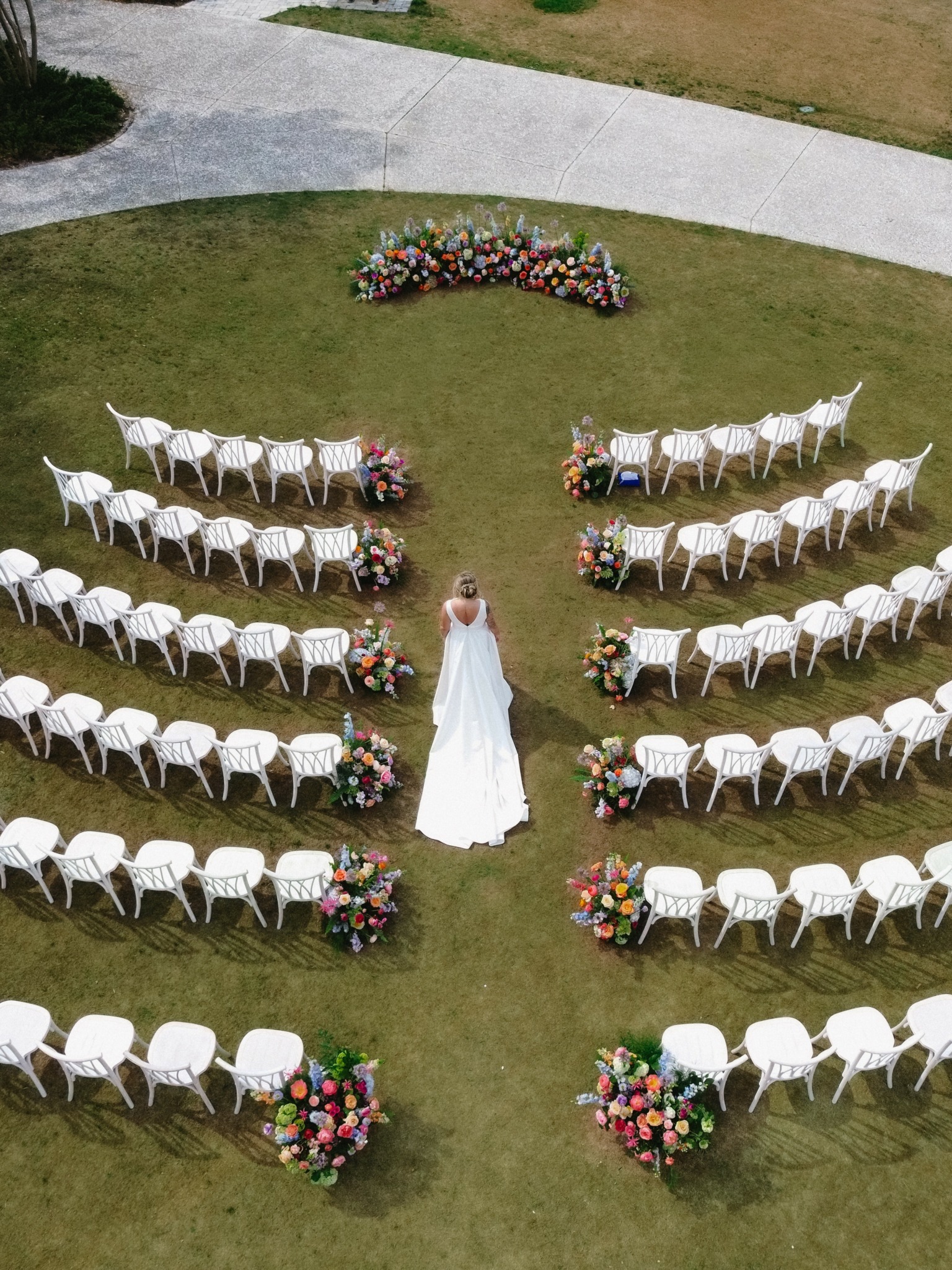 A bride in a white dress stands in the center of an outdoor wedding setup with white chairs and colorful flower arrangements, viewed from above.