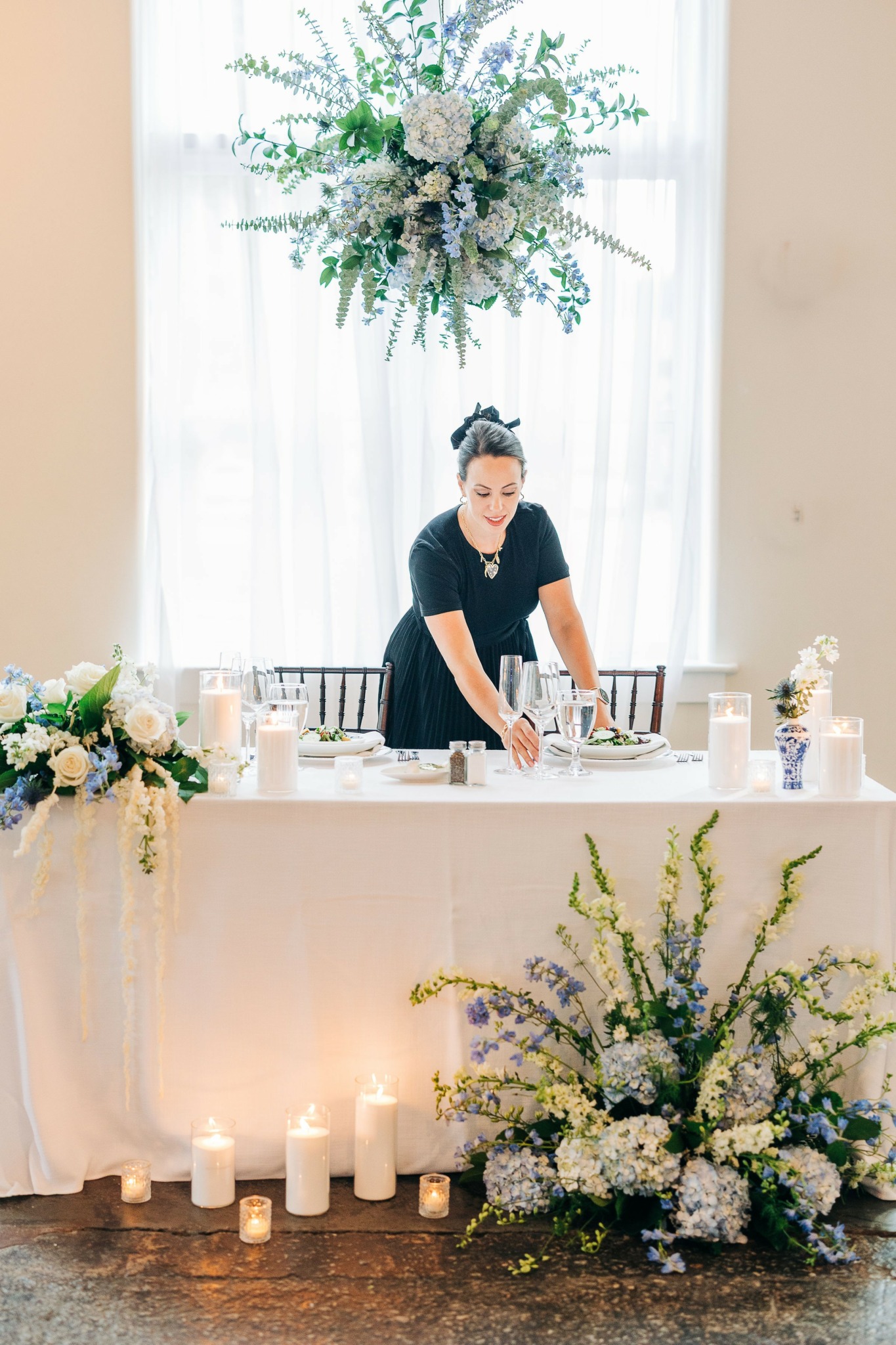 Woman arranging items on a table with floral decorations and candles, in front of a window with a large floral arrangement above.