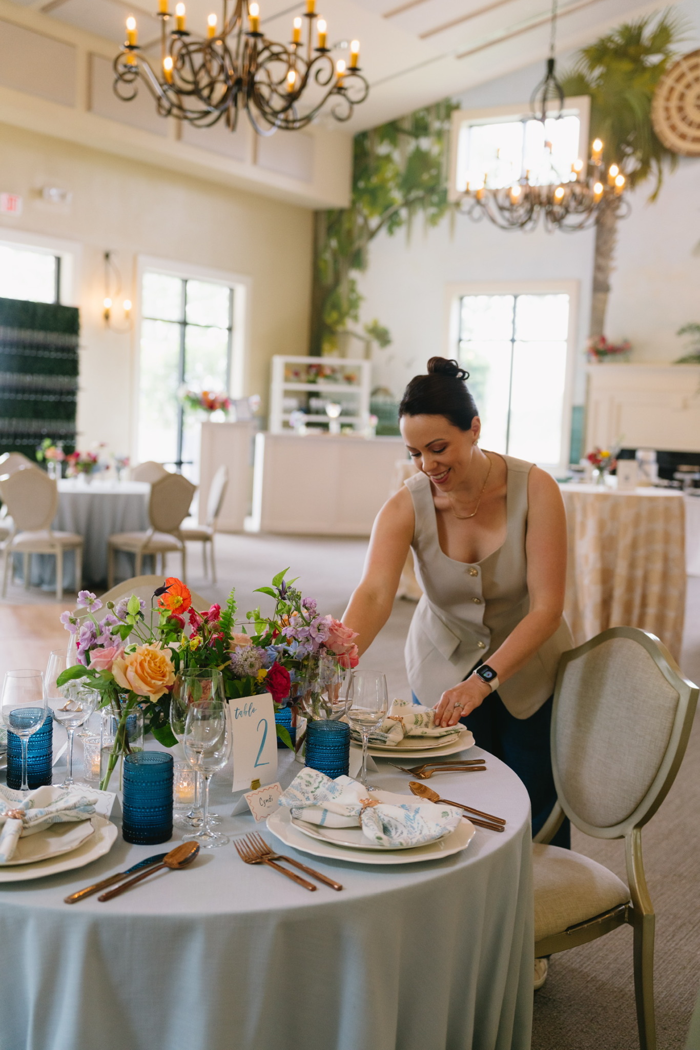 Woman arranging table settings in a decorated dining room with chandeliers and floral arrangements.