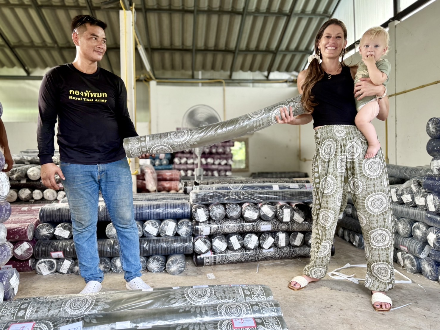 Three people in a warehouse with stacked metal pipes, a woman holding a child, and a man holding a pipe, smiling.