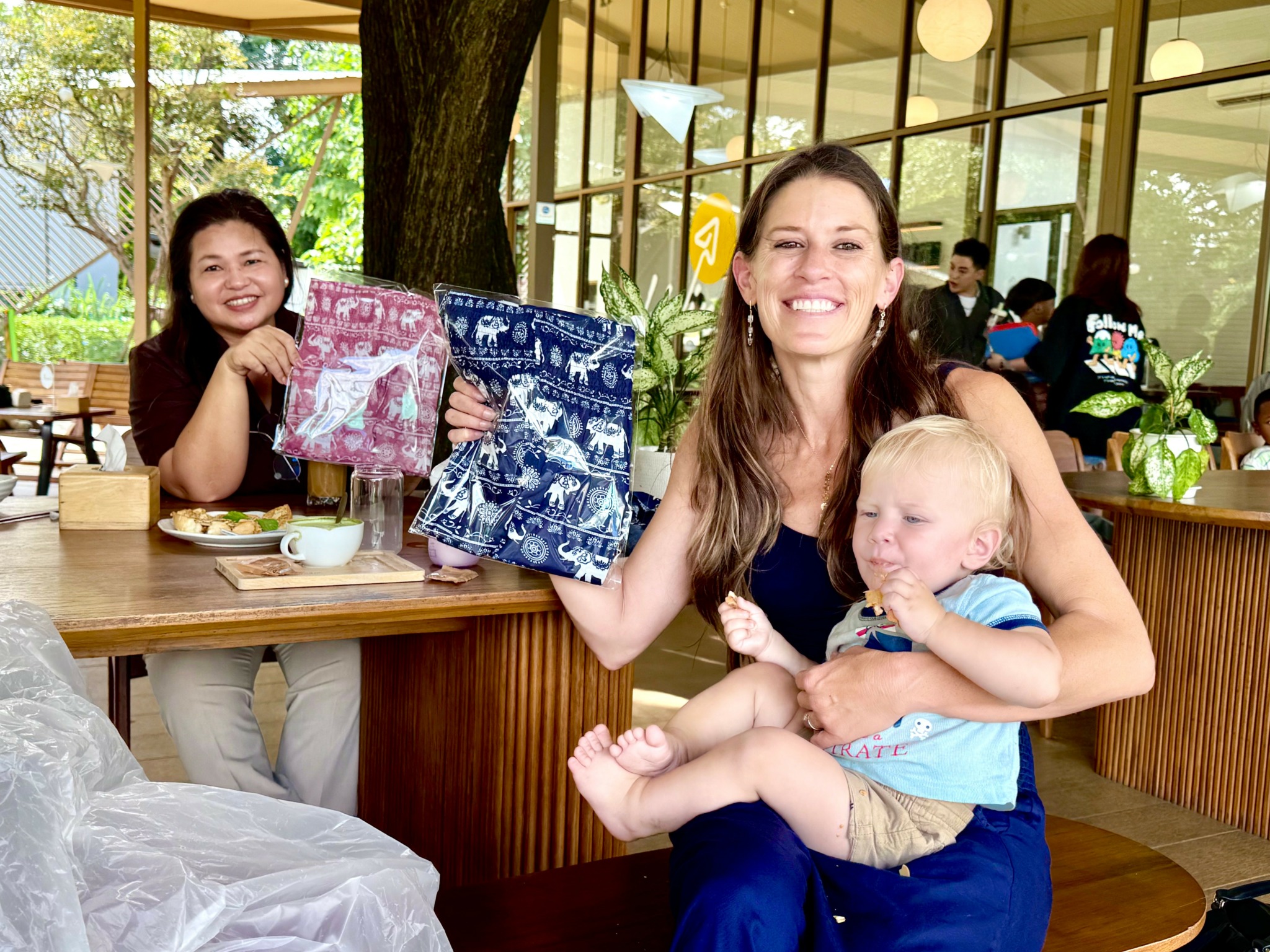 Two women and a child sitting at a table in a cafe, smiling and holding colorful books, with large windows and trees outside.