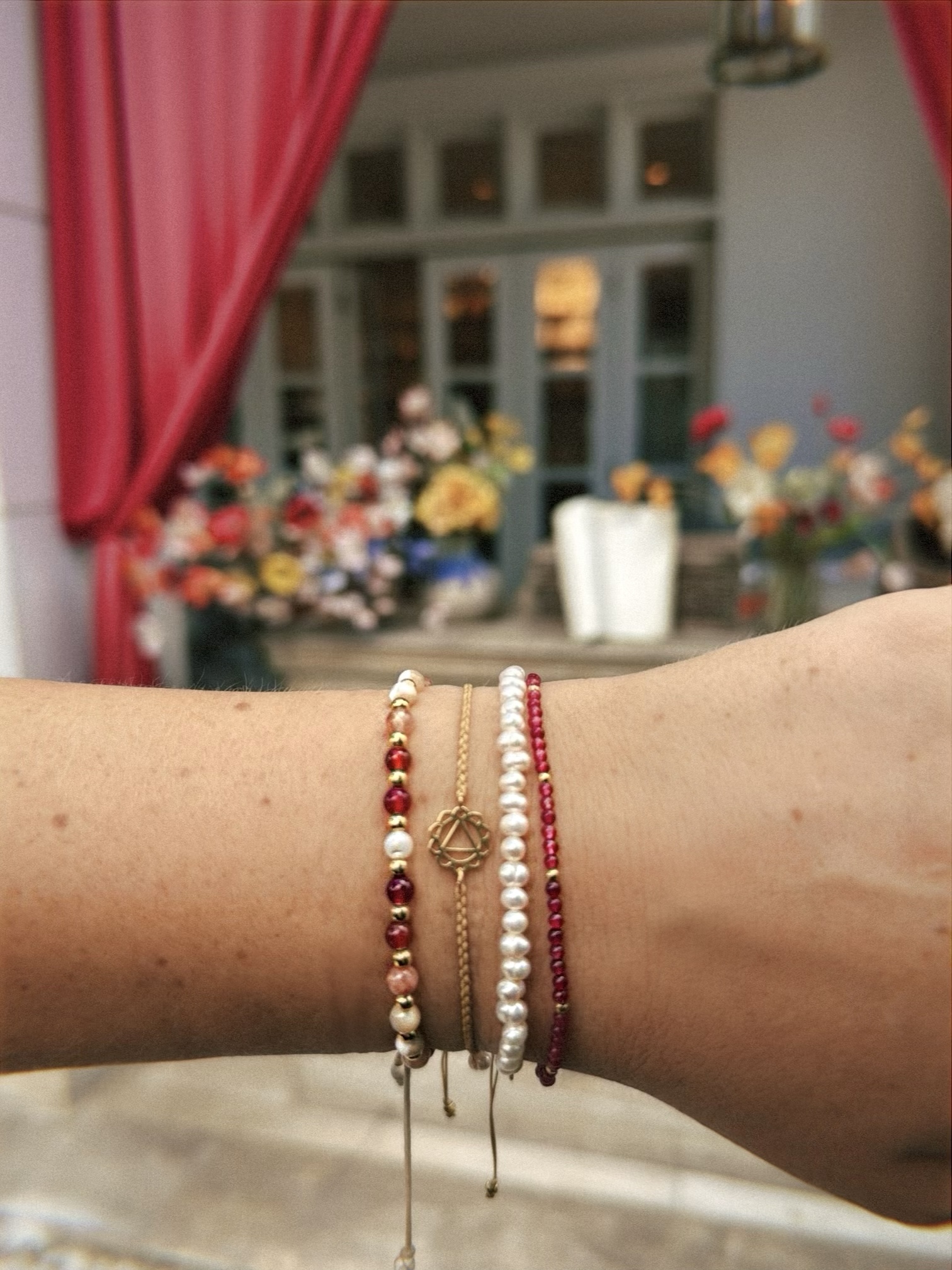 Person's wrist with multiple bracelets, background of a room with red curtains and a table with flowers.