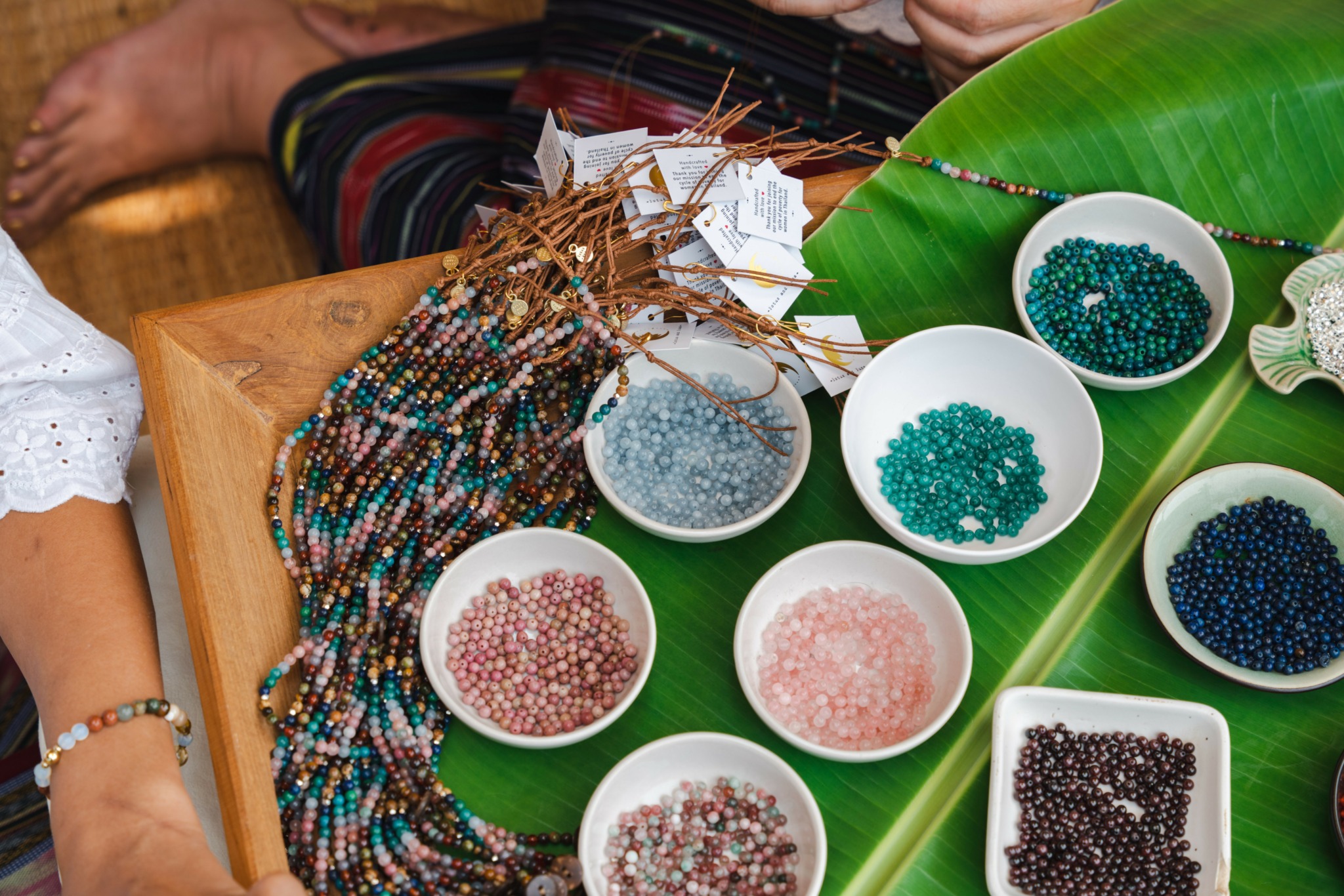 Beads and jewelry-making supplies on a wooden table with green leaves, bowls of beads, and a person's arm visible.