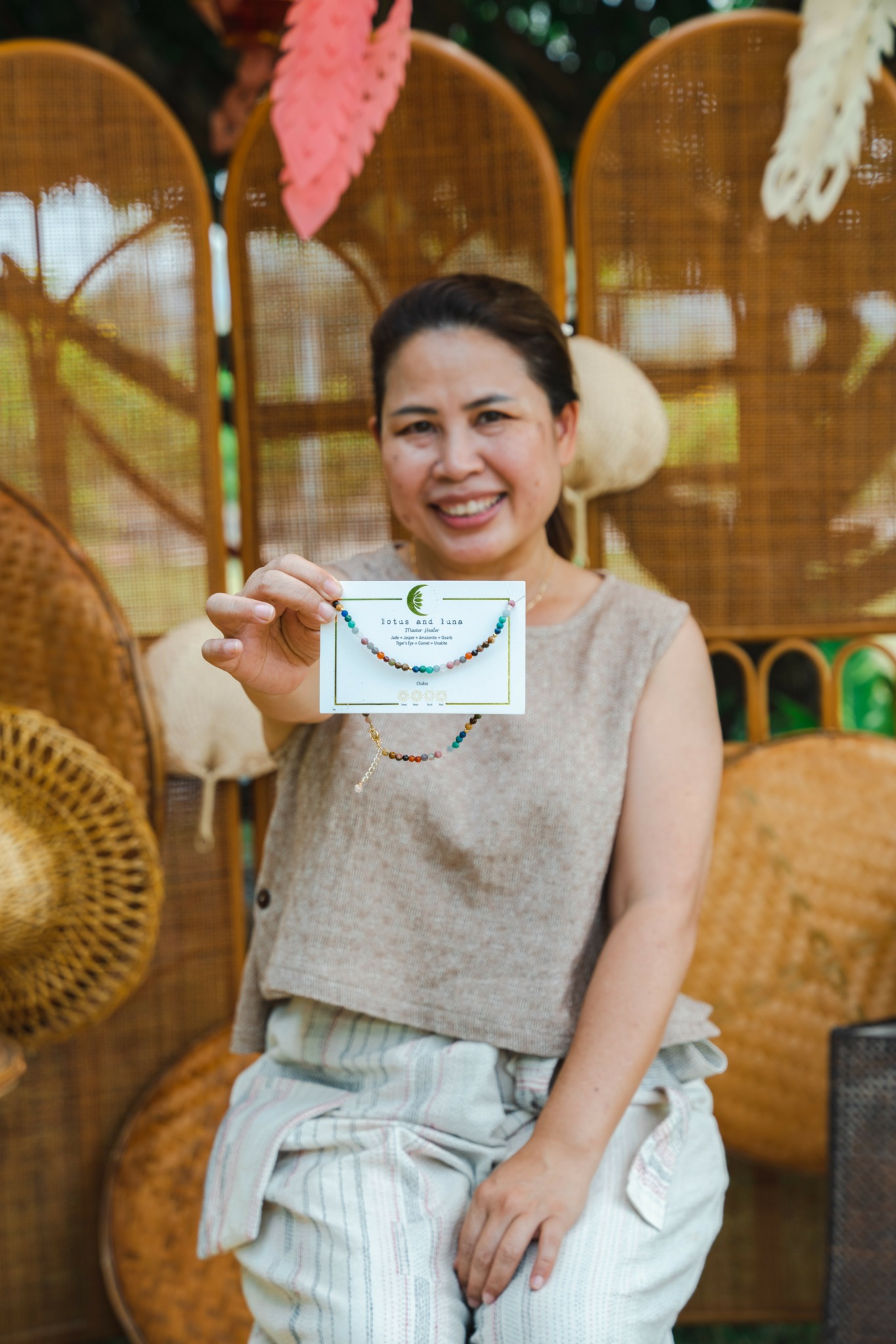 Smiling woman holding a certificate, sitting in front of woven chairs and colorful decorations.