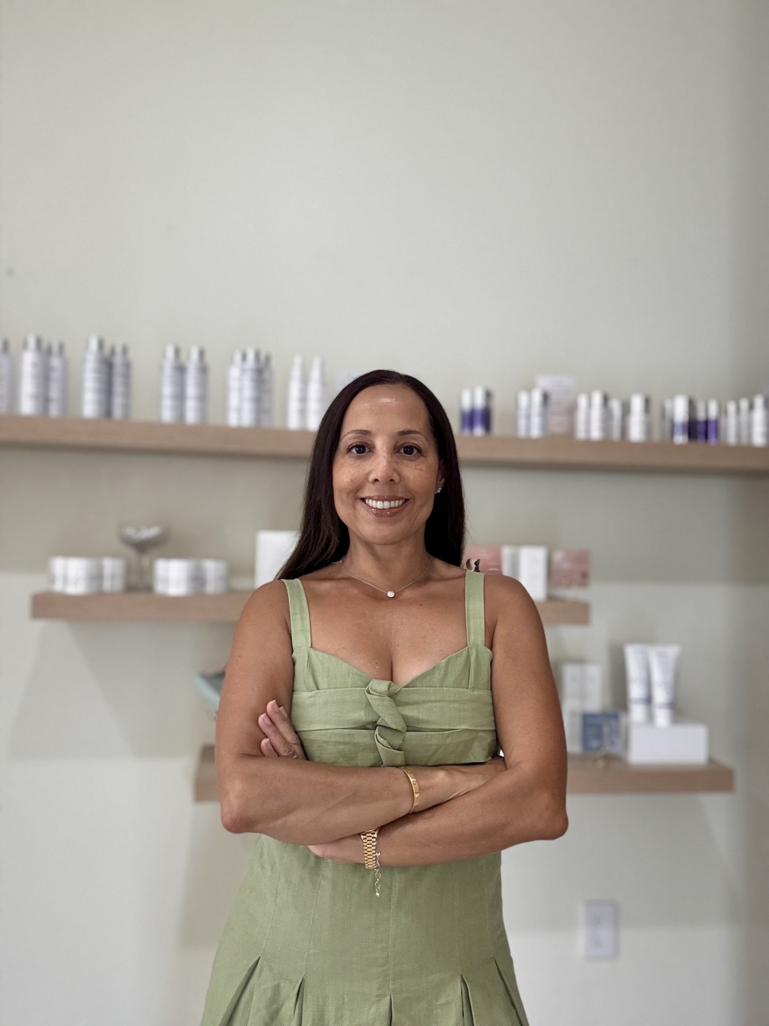 Smiling woman with long dark hair in a green dress standing with arms crossed in front of shelves with bottles and containers.