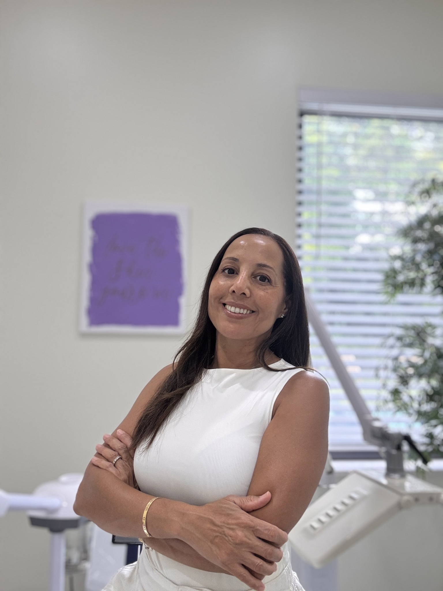Smiling woman with long dark hair wearing a white sleeveless top, arms crossed, in a room with a window and a purple artwork.