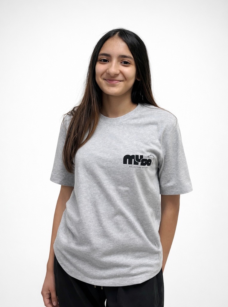 Young woman with long dark hair smiling, wearing a gray T-shirt and black pants, standing against a white background.