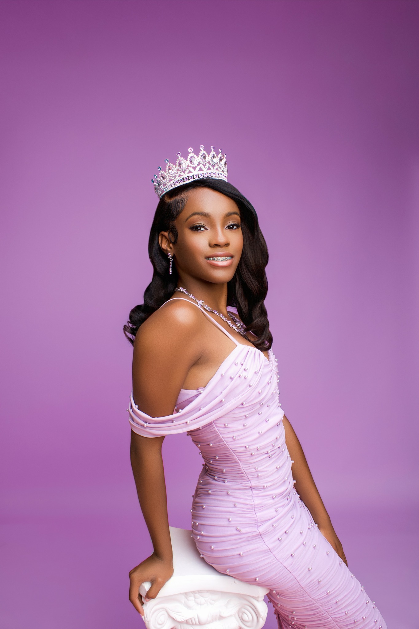 Young woman wearing a crown and a pink dress, posing against a purple background.