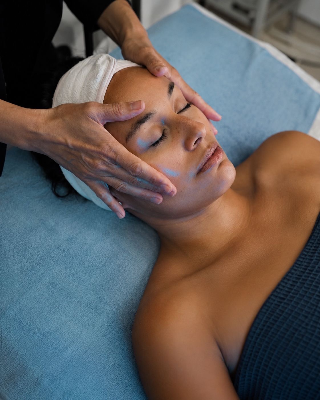 Person receiving facial treatment with eyes closed, lying on a blue surface, with hands gently massaging face, head wrapped in towel.