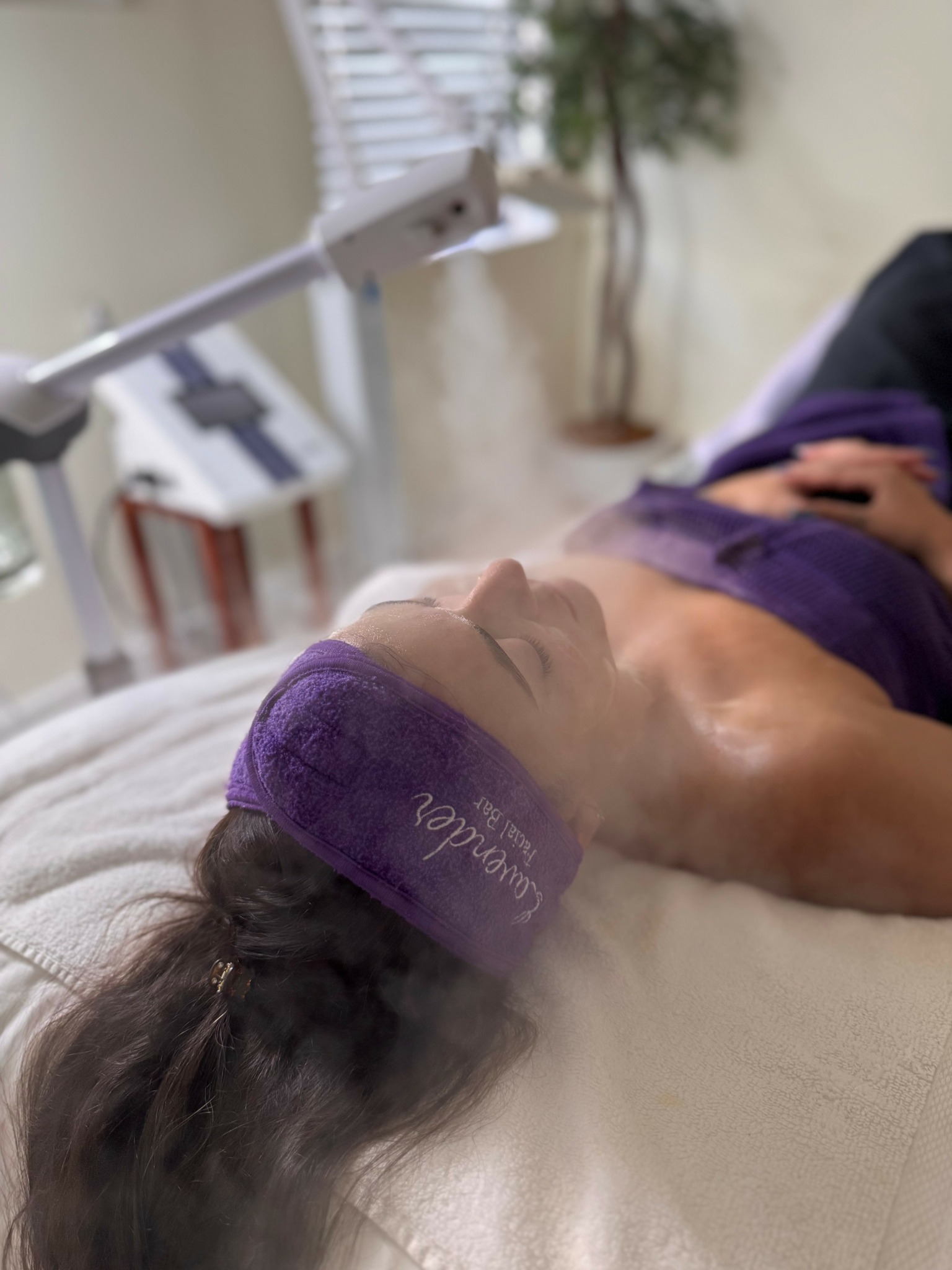 Person lying on a treatment bed with a purple headband, eyes closed, in a clinical setting with medical equipment nearby.