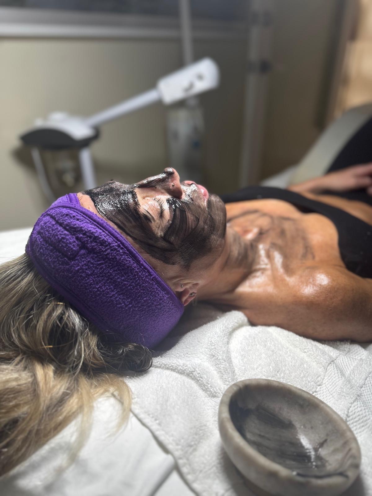 A person with a purple headband receives a facial treatment, lying on a bed with a bowl nearby.