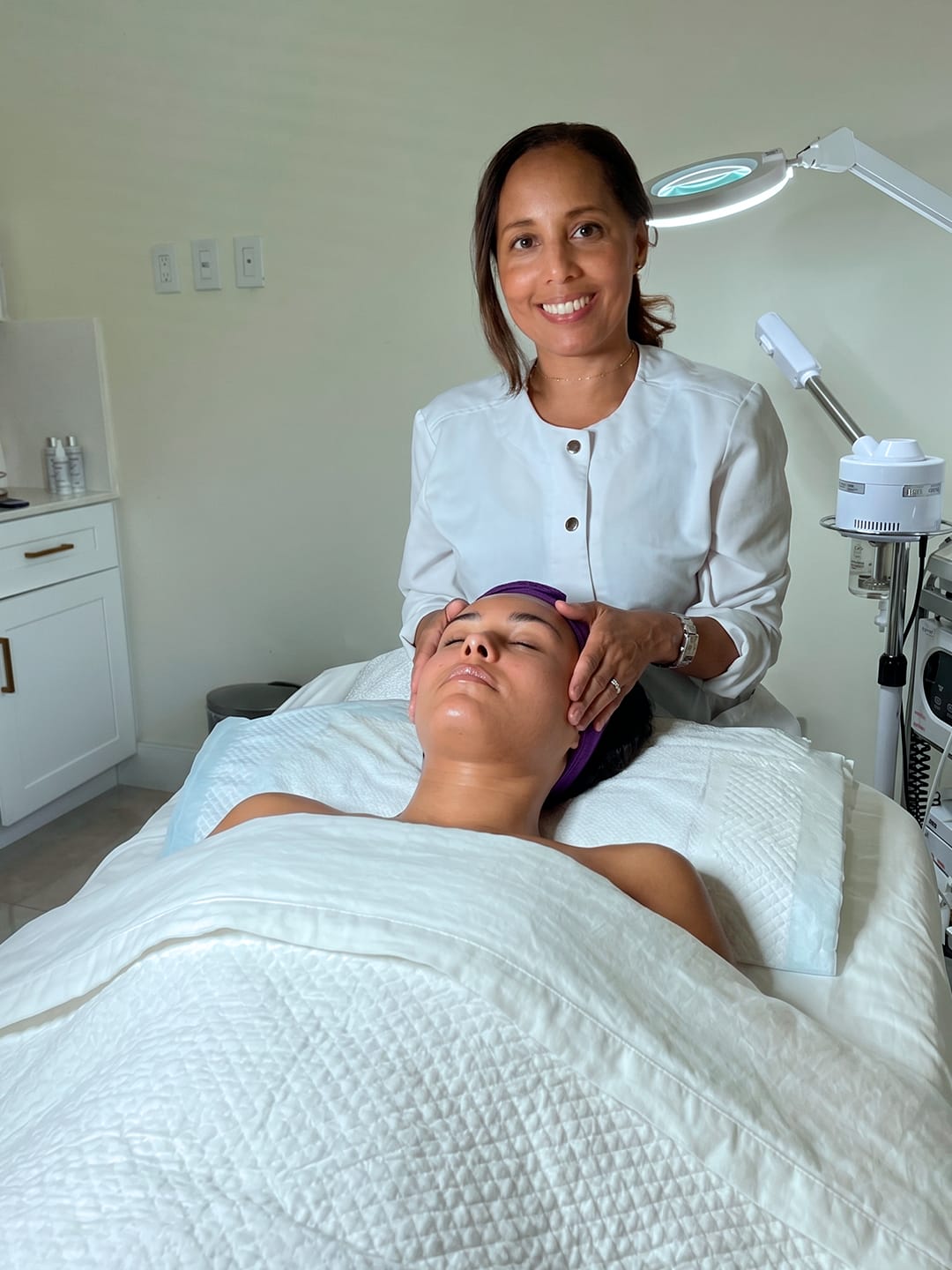 A woman lying on a bed with her eyes closed, covered with a white blanket, in a medical setting. A smiling woman stands beside her, holding her hand. Medical equipment is visible in the background.