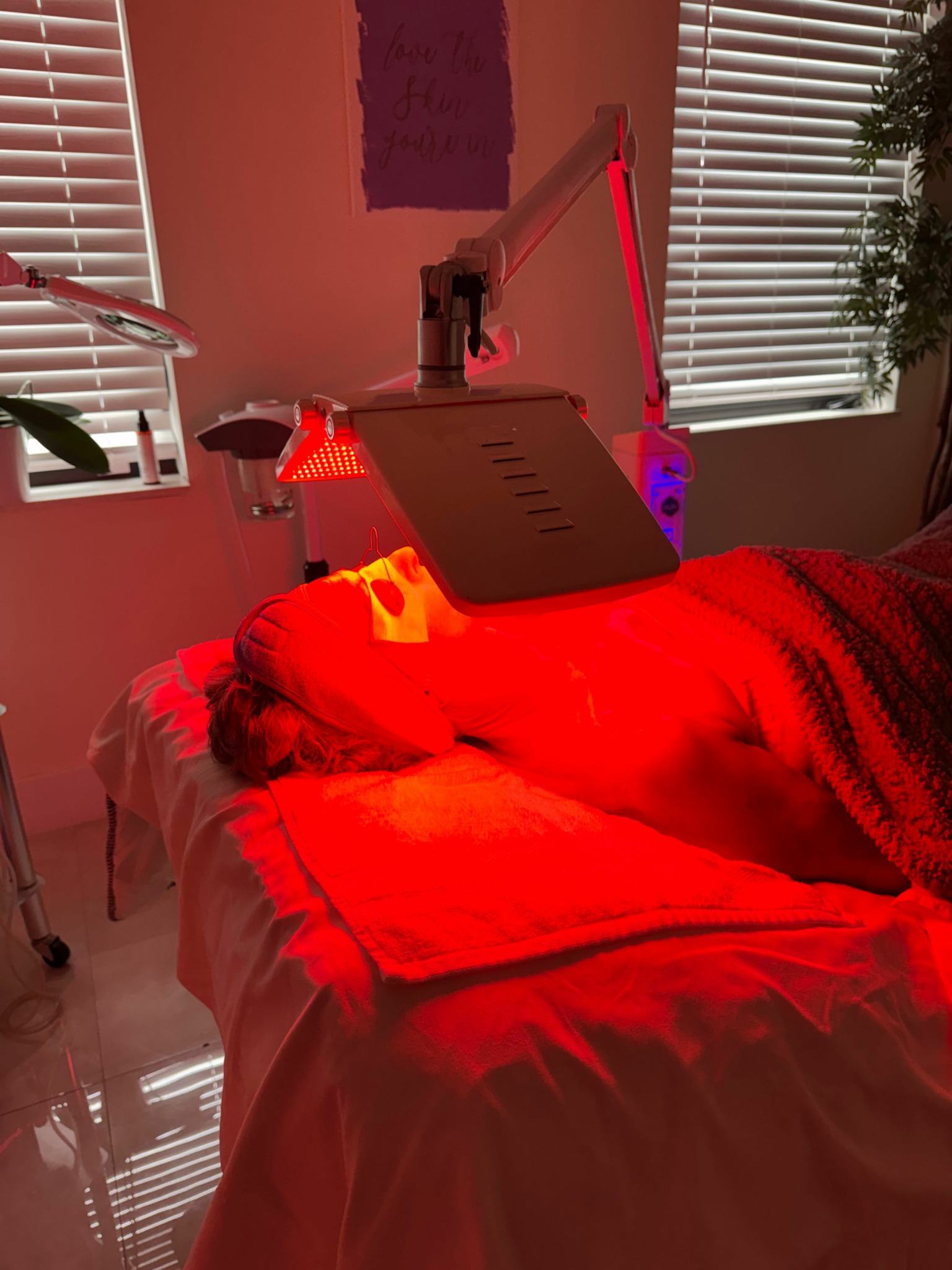 A room with a bed, a laptop, and a window with blinds, illuminated by red light, with a radiator on the wall.