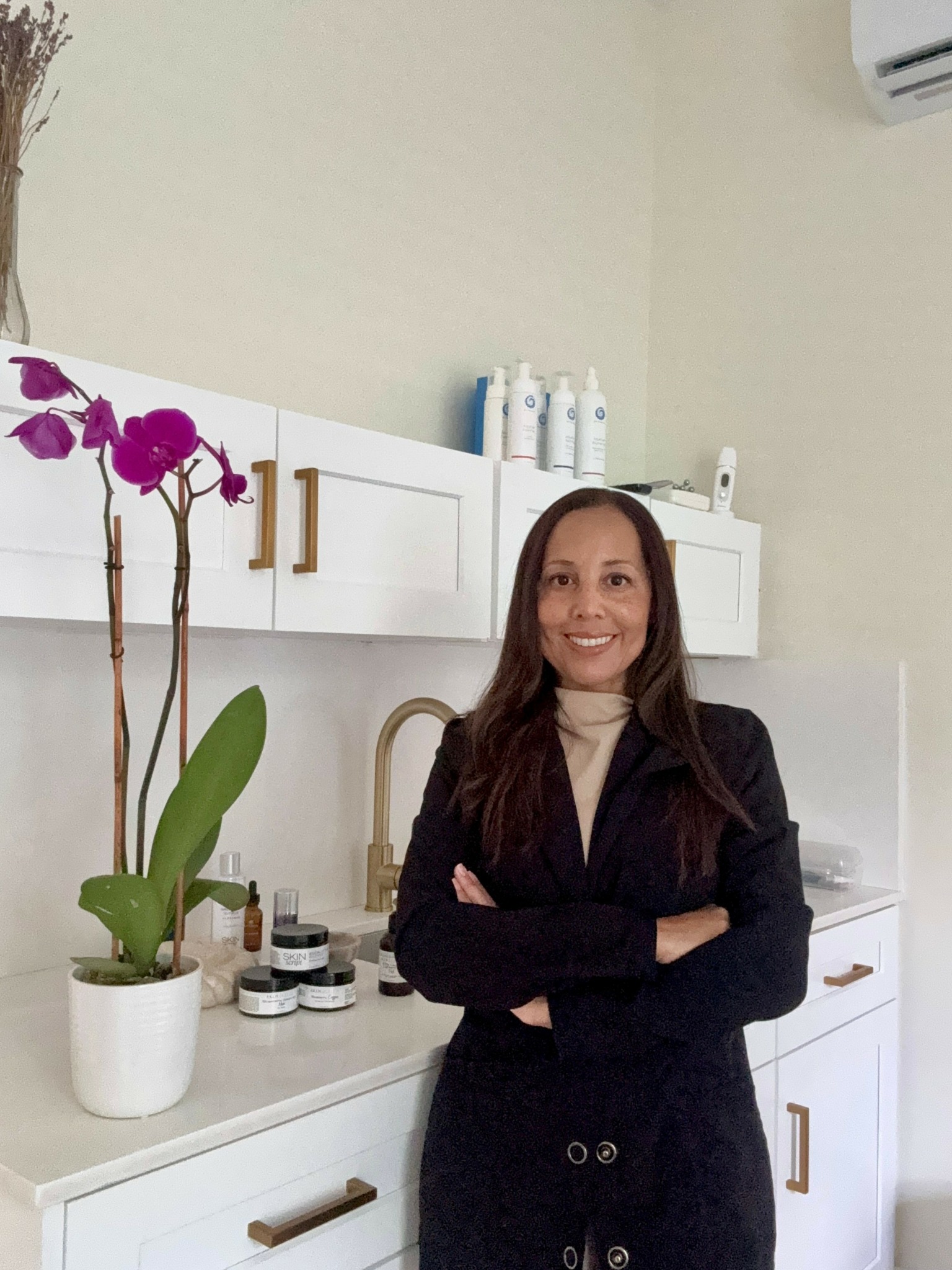 Woman with long hair smiling, standing in a kitchen with white cabinets and a potted plant.