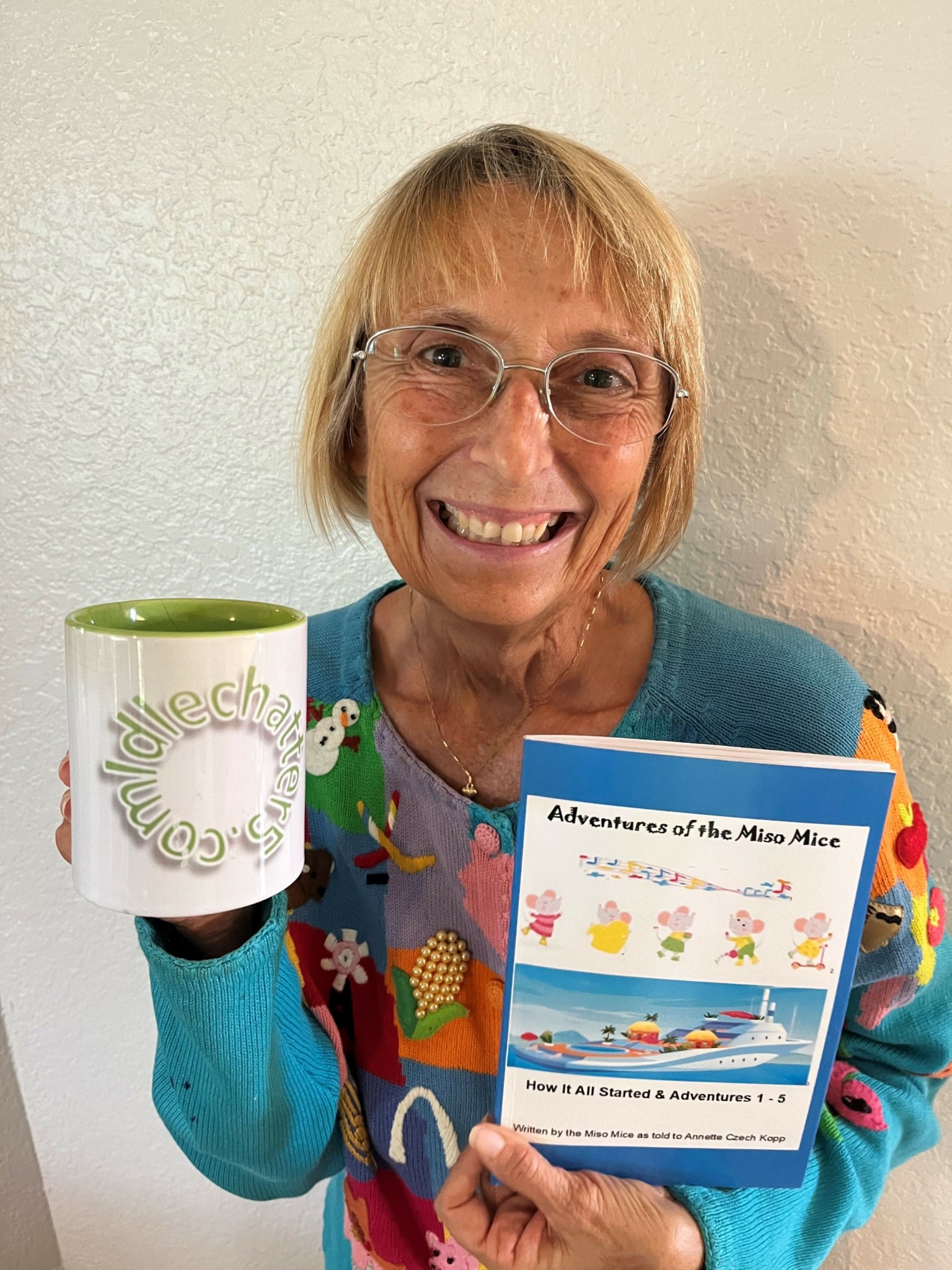 Older woman with glasses smiling, holding a colorful booklet and a white mug with a green interior, against a plain wall.