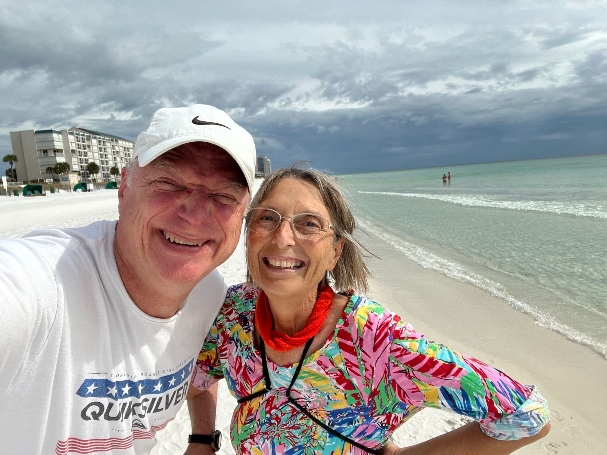 Two people smiling on a beach with ocean waves and cloudy sky in the background.
