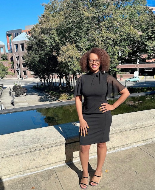 Woman with curly hair in a black dress and heels standing outdoors near a stone railing, trees, and buildings.