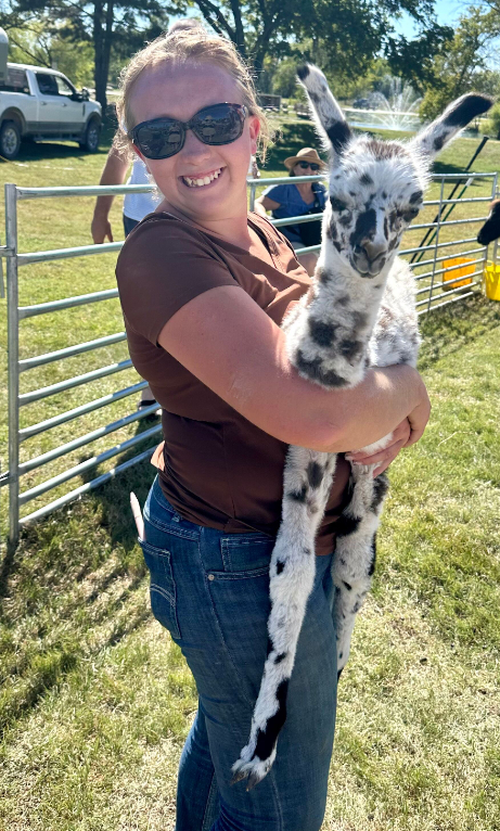 Woman holding a spotted baby giraffe outdoors, smiling, wearing sunglasses and a brown shirt.
