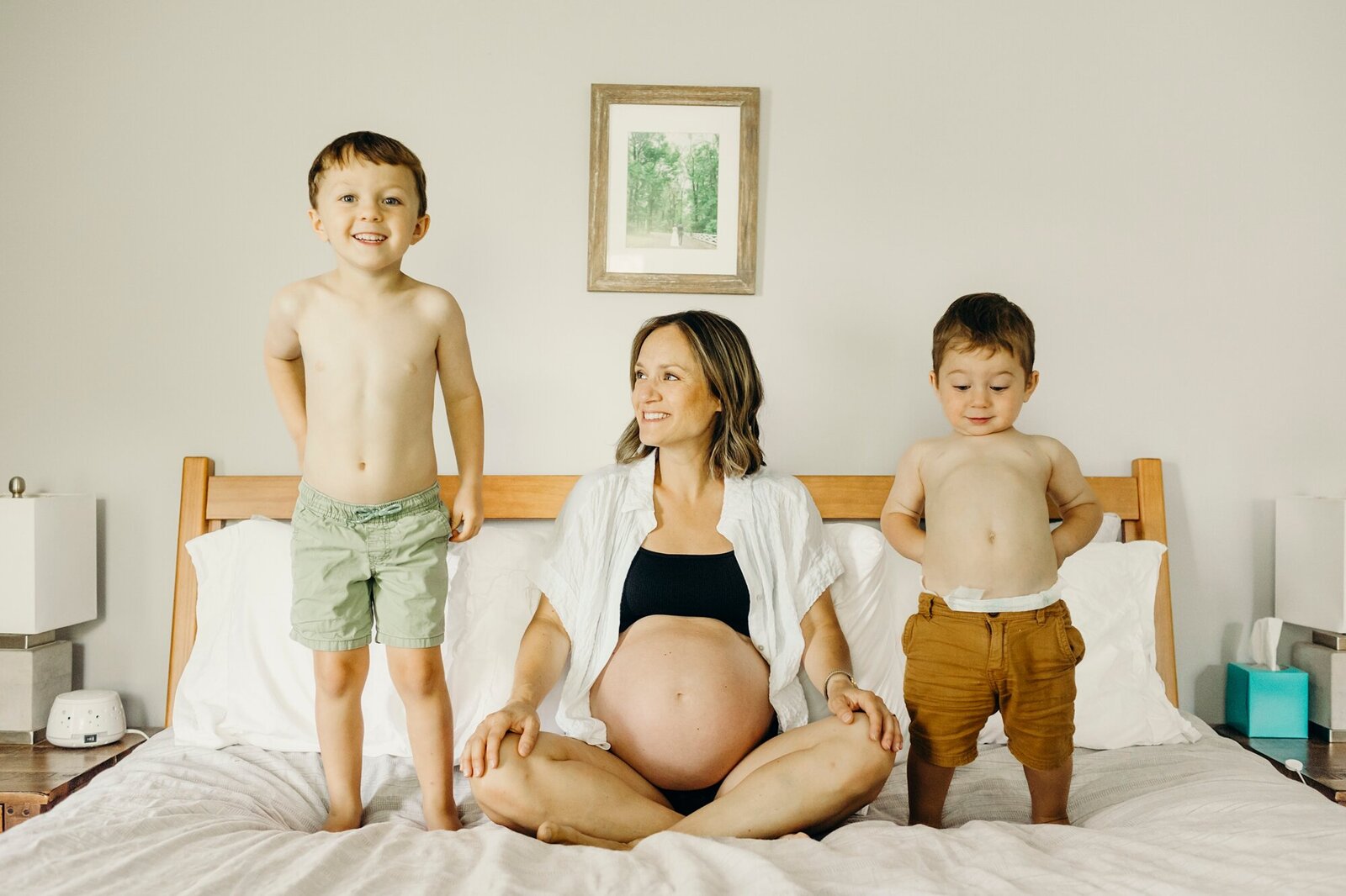 Pregnant woman sitting on bed with two children, one standing and one sitting, in a bedroom with framed picture on wall
