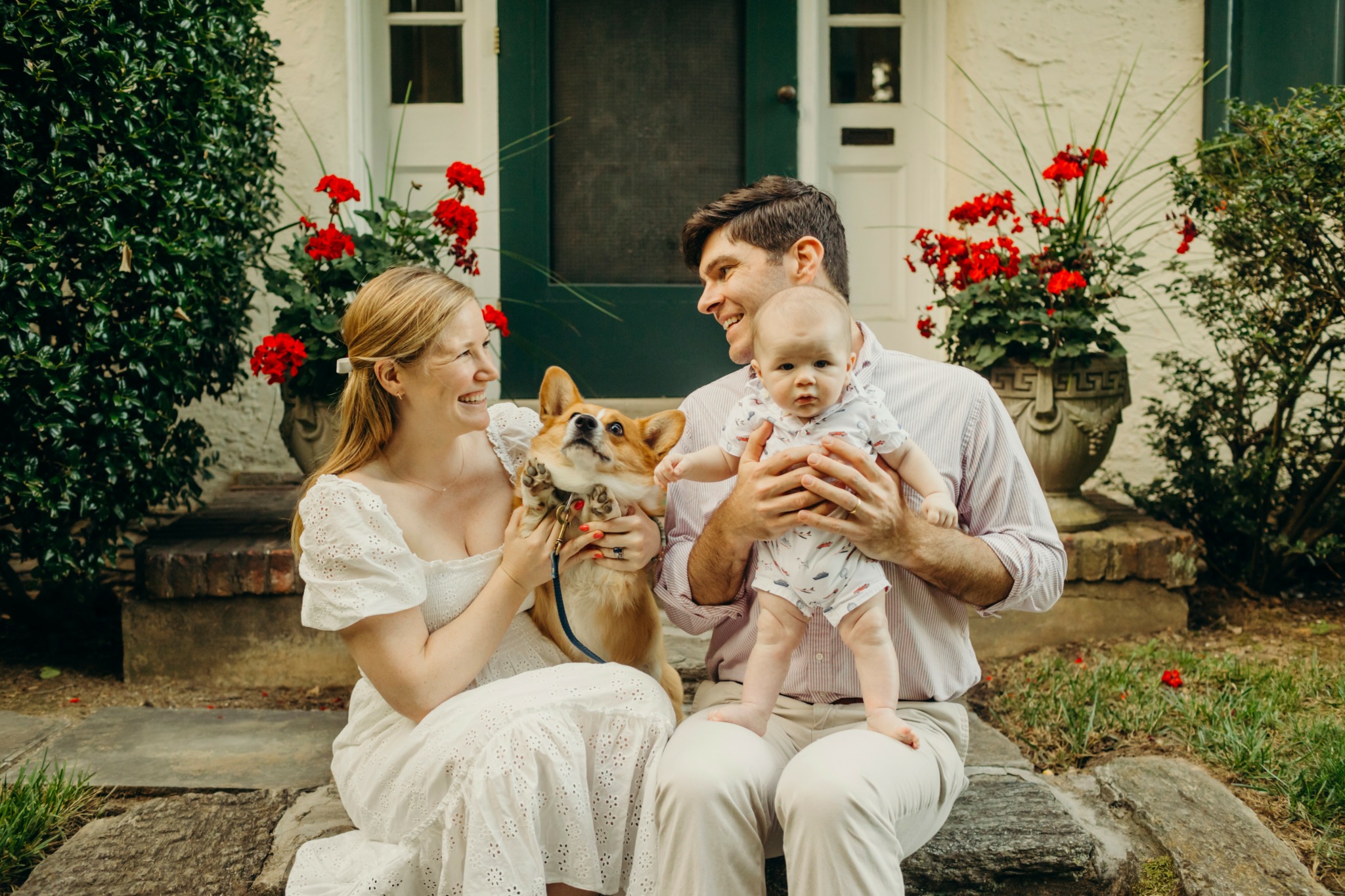 Family with a dog sitting on steps outside a house with red flowers, smiling and playing together.