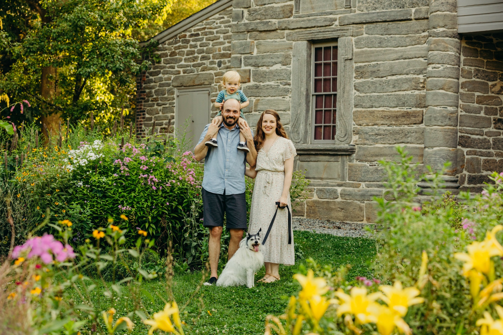 Family of three with a dog standing outdoors near a stone building, surrounded by colorful flowers and greenery.