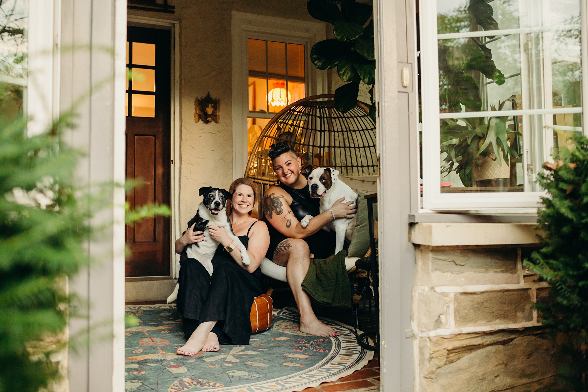 Two women and a man sitting on a porch with two dogs, smiling and holding the dogs, surrounded by plants and a cozy interior.