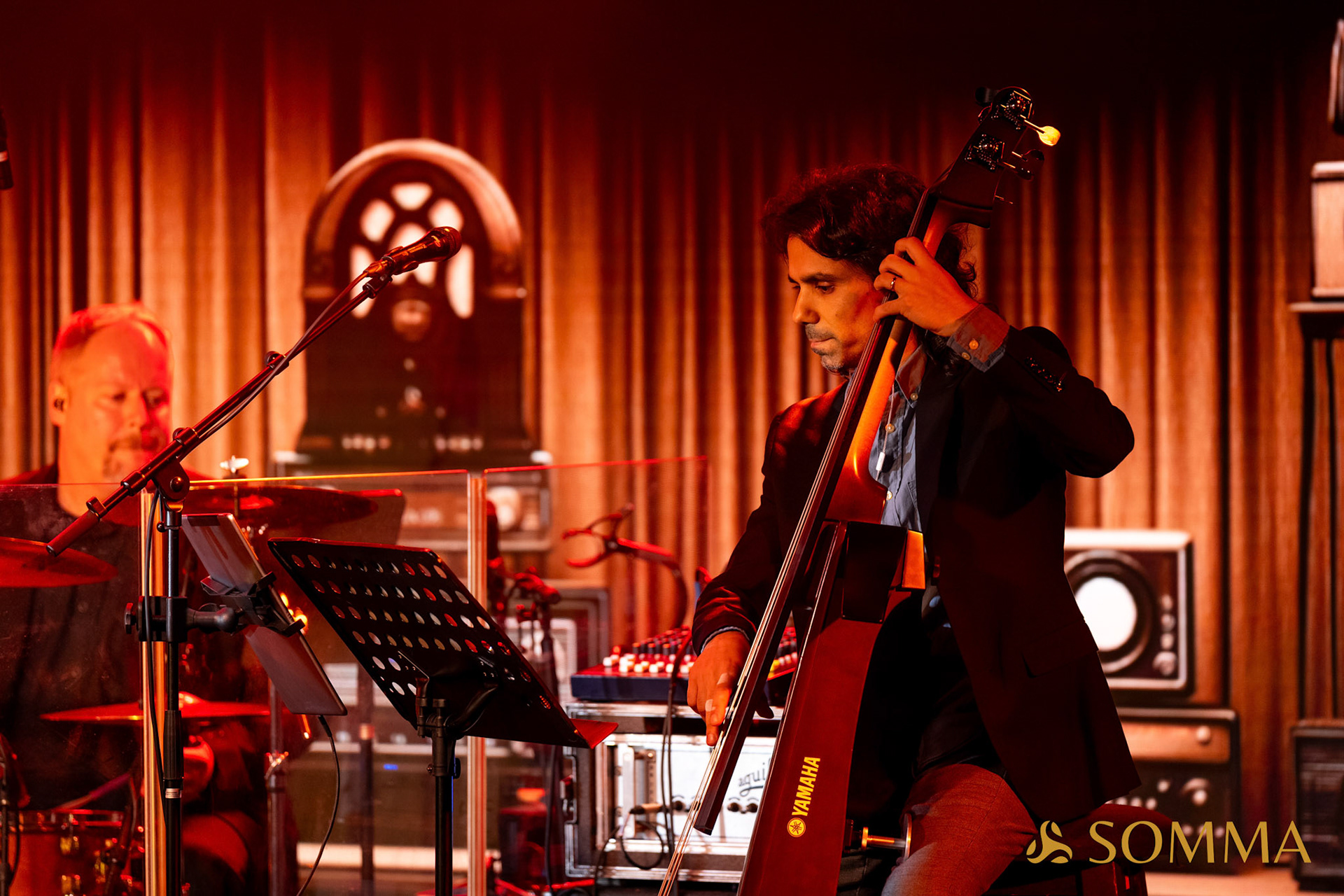 Two musicians perform on stage with a cello and keyboard, warm lighting, and a curtain backdrop.