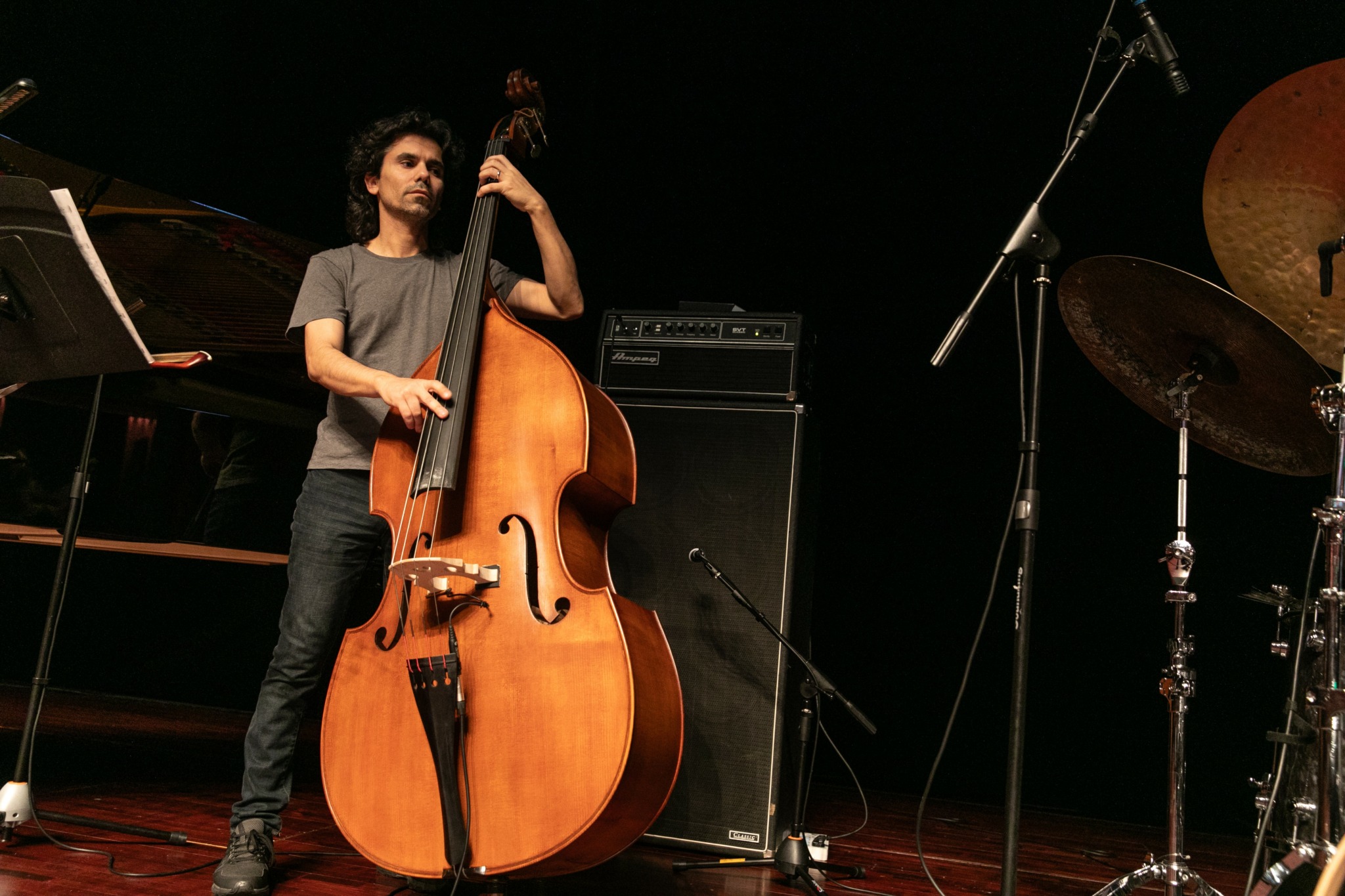 Young man playing double bass on stage with musical equipment, microphone, and drum set in background.