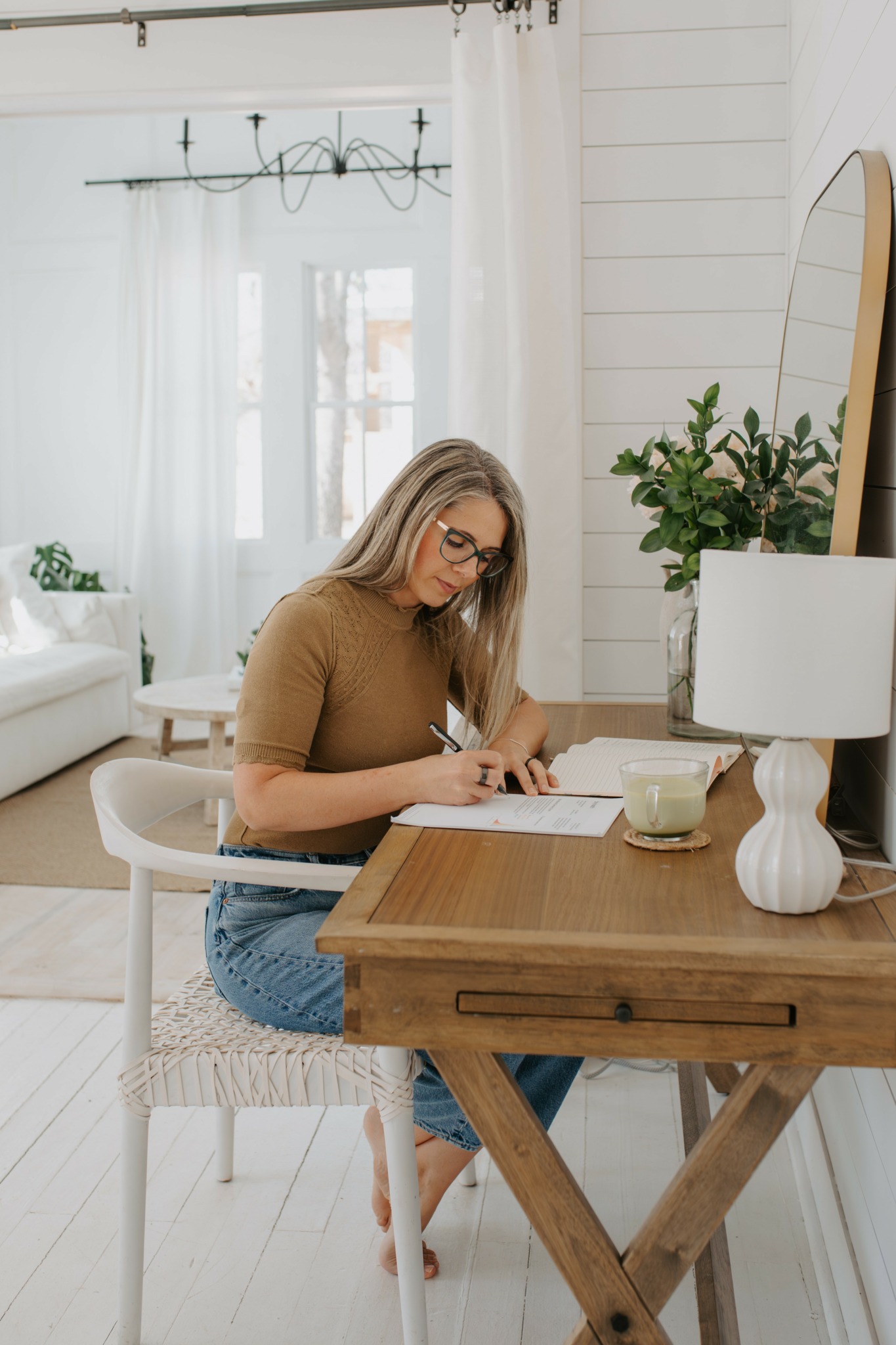 Woman sitting at a wooden table writing in a notebook, with a cup and a lamp, in a bright room with white walls and window.