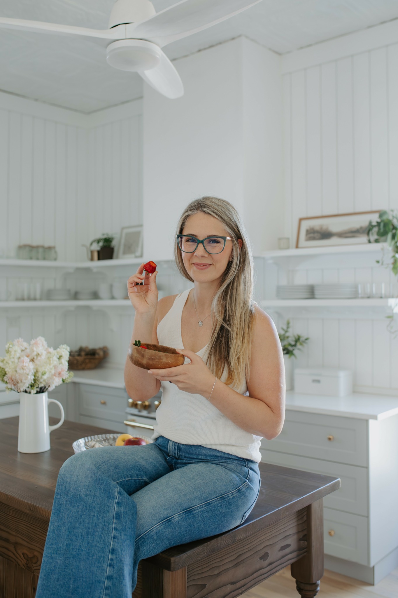 Young woman with glasses sitting on a wooden table in a bright kitchen, holding a strawberry and a bowl.