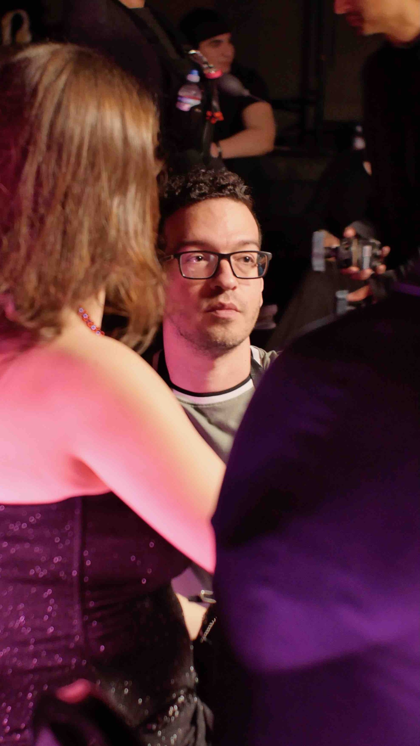 A man with glasses and curly hair looks at a woman with shoulder-length hair, in a dimly lit setting.