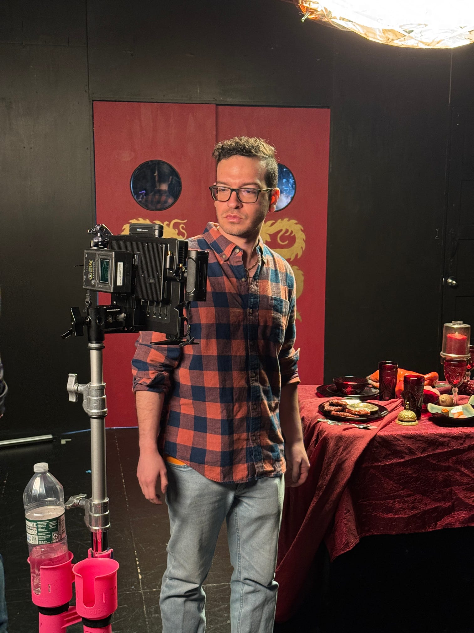 Man with glasses in plaid shirt standing next to camera and table with food and drinks, black background, red decorative panel.